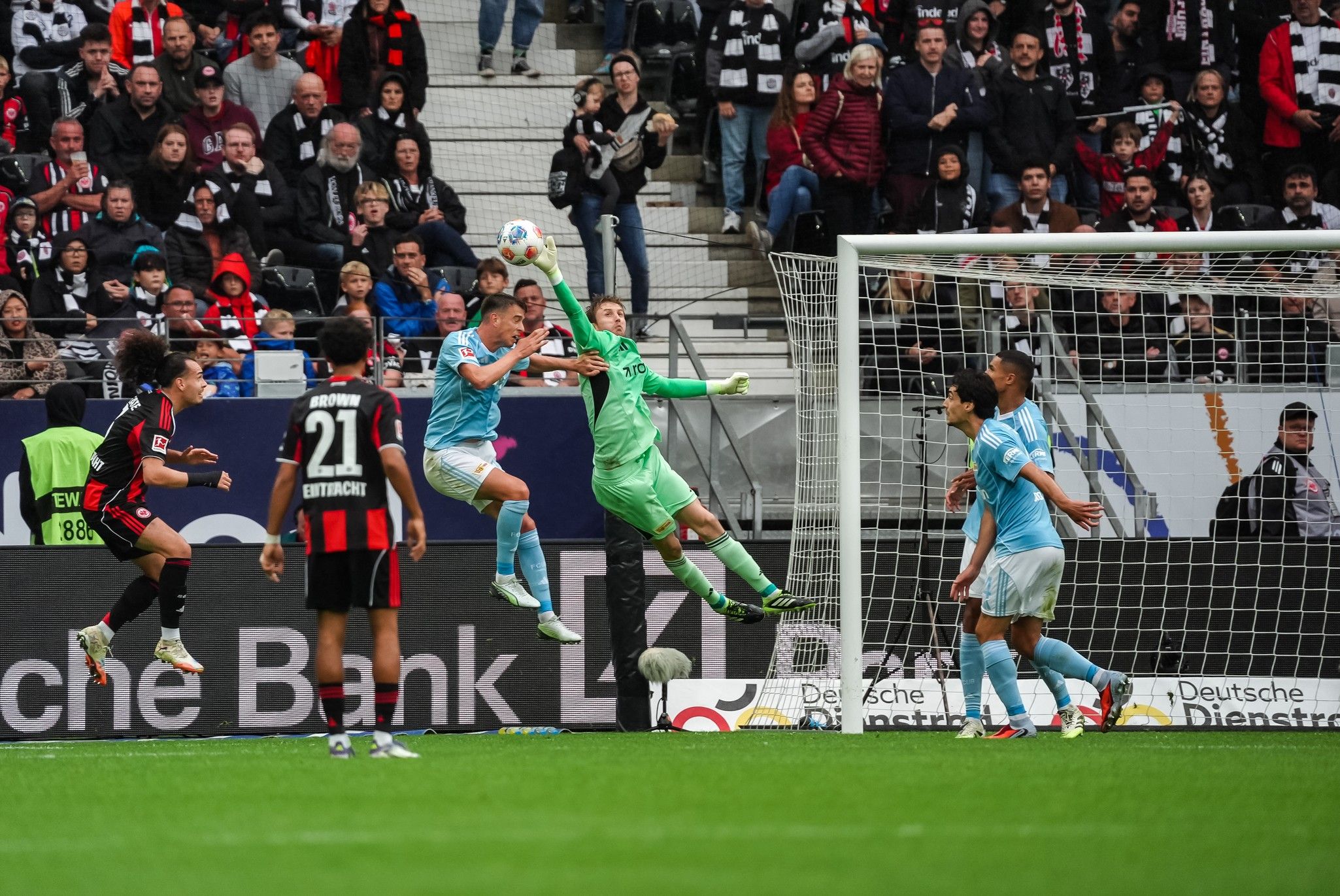 Zwei Fußballspieler kämpfen um den Ball, während der Torwart einen Schuss abwehrt, umgeben von jubelnden Fans.