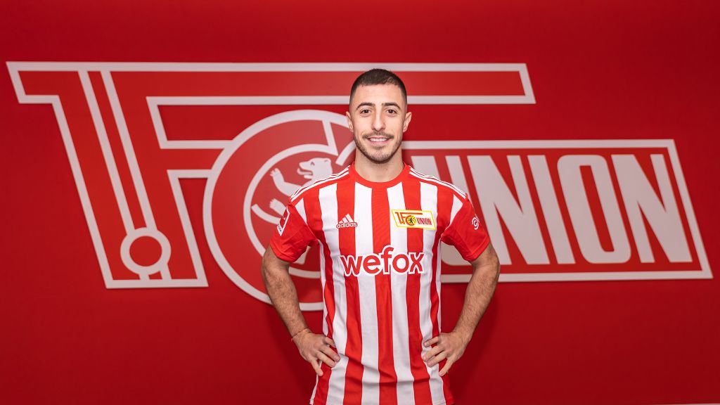 Ein Fußballspieler in einem rot-weiß gestreiften Trikot steht vor einem roten Hintergrund mit dem Logo des FC Union Berlin.