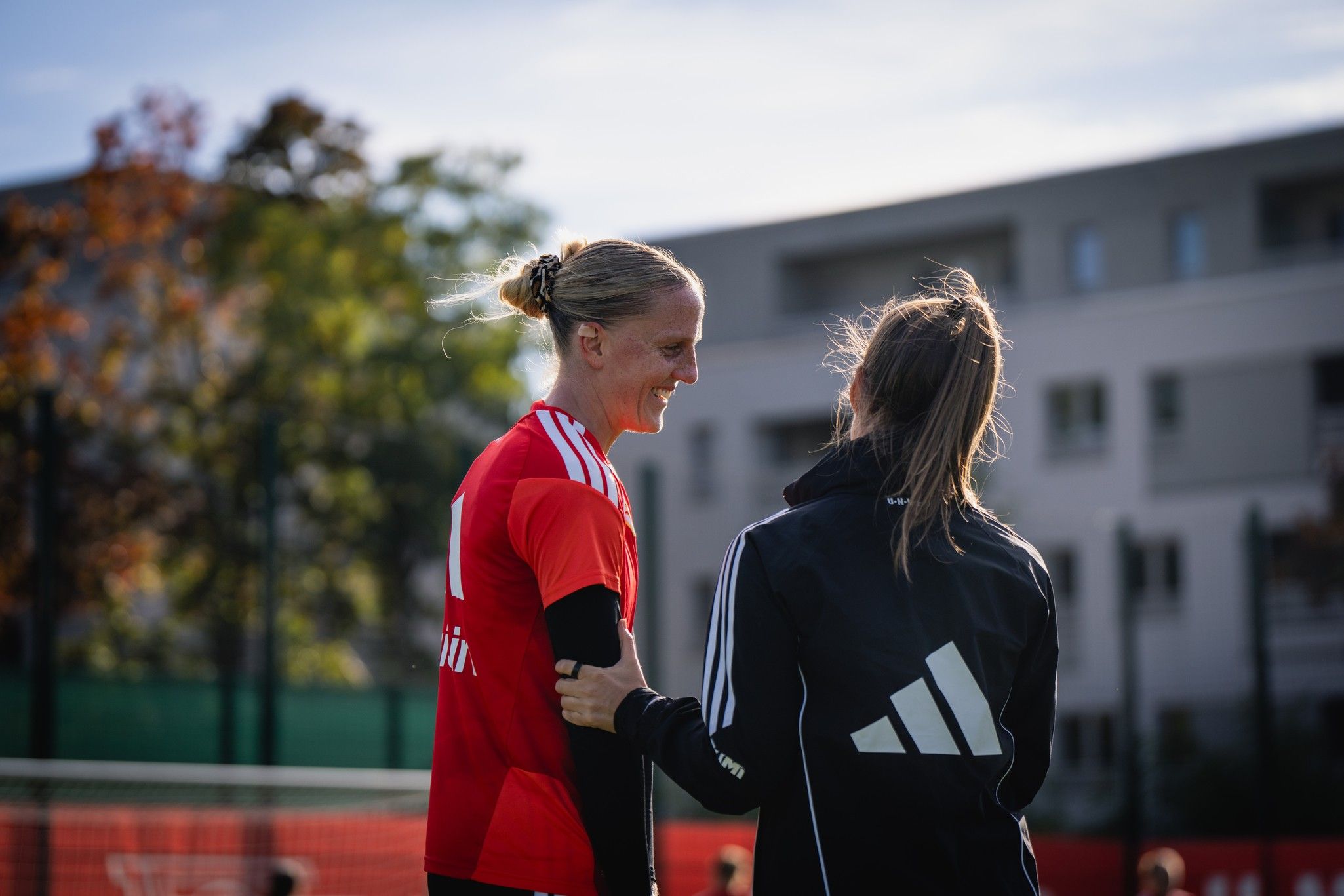 Zwei Frauen im Gespräch auf einem Fußballplatz, eine trägt ein rotes Trikot, die andere eine schwarze Jacke mit weißem Logo.