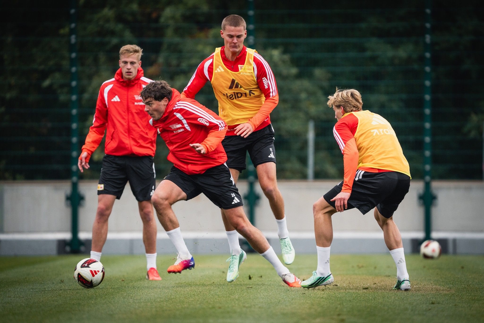 Vier Fußballspieler in Trainingsanzügen führen ein Übungsspiel auf einem Rasenfeld durch.