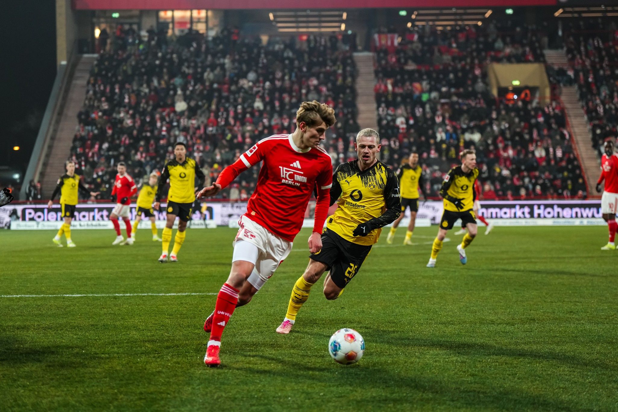 Ein Fußballspieler in roter Kleidung dribbelt den Ball, während ein gegnerischer Spieler in gelber Kleidung ihn verfolgt. Zuschauer im Hintergrund.
