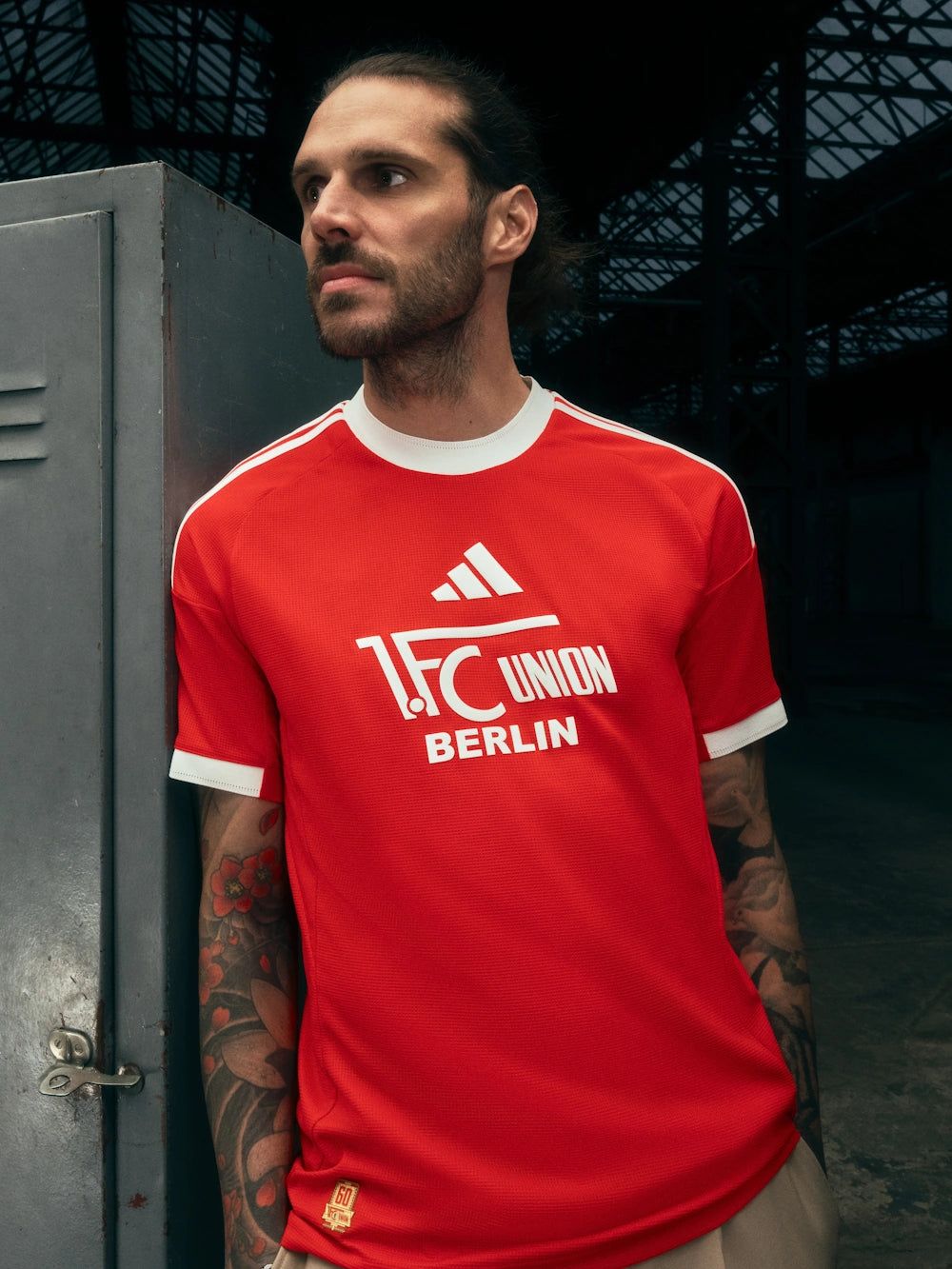 Ein Mann in einem roten T-Shirt mit dem Logo von 1. FC Union Berlin steht an einer Wand in einer industriellen Umgebung.