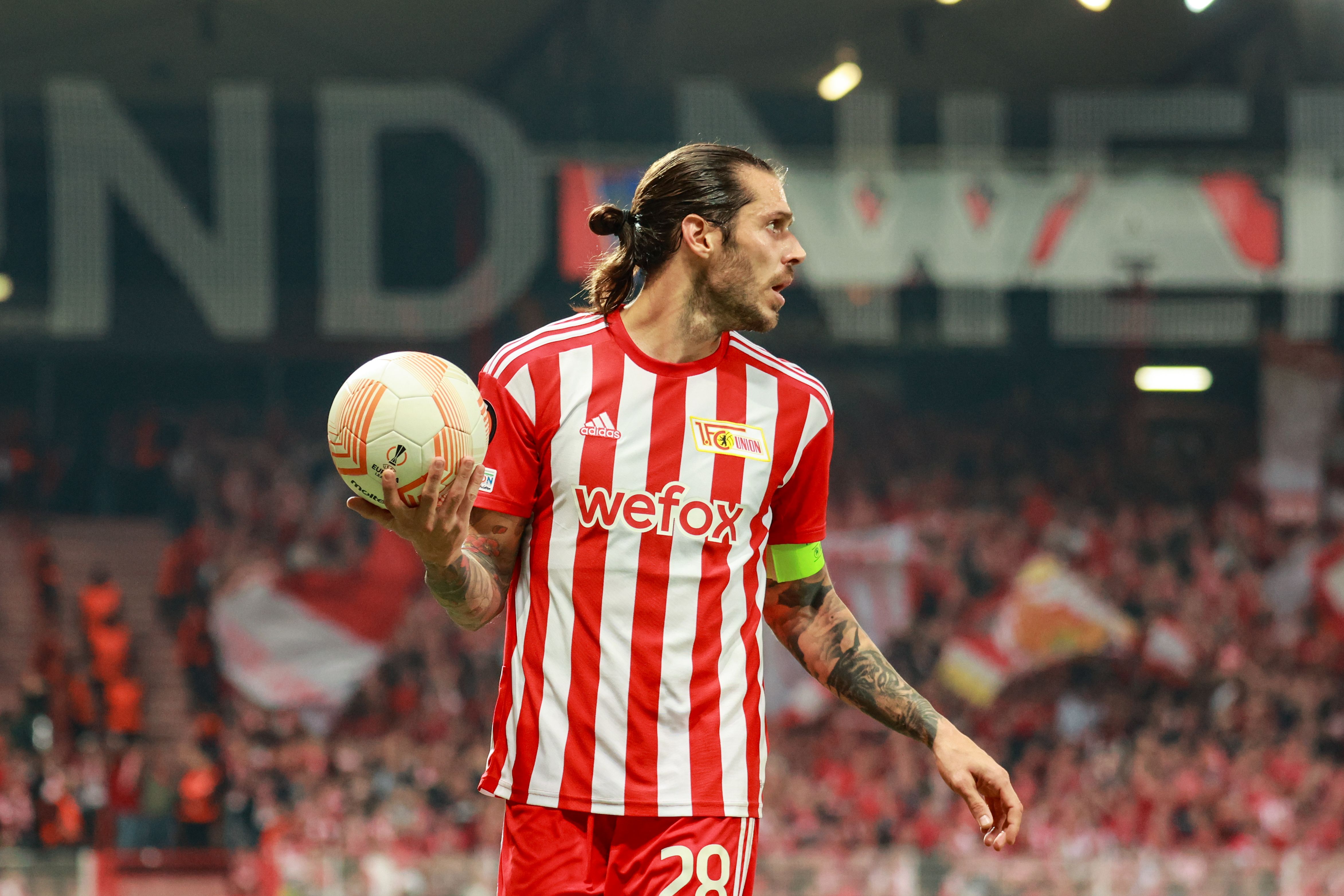 Ein Fußballspieler in rot-weißem Trikot hält einen Ball und schaut auf das Publikum, während Fans im Hintergrund jubeln.