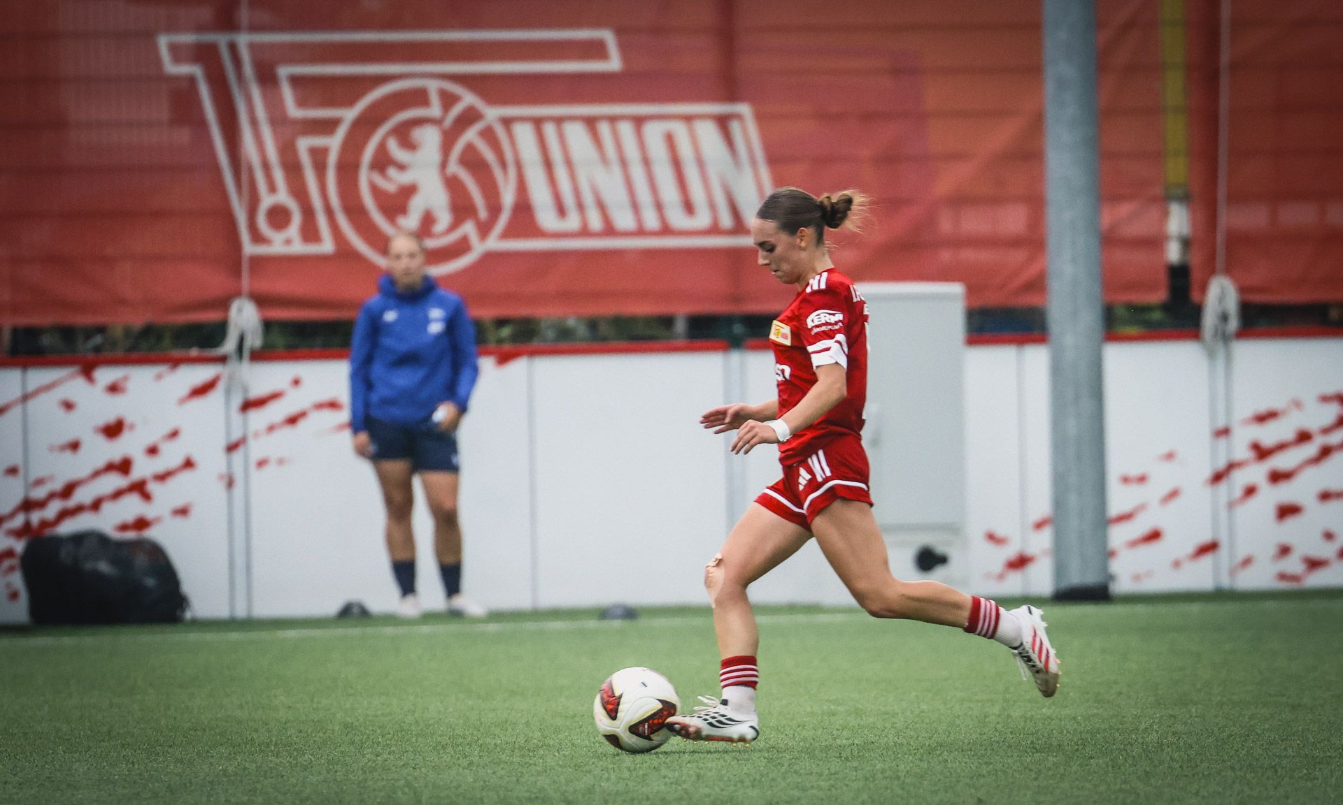 Eine Fußballspielerin in rot spielt den Ball auf einem Trainingplatz, im Hintergrund steht eine Trainerin.
