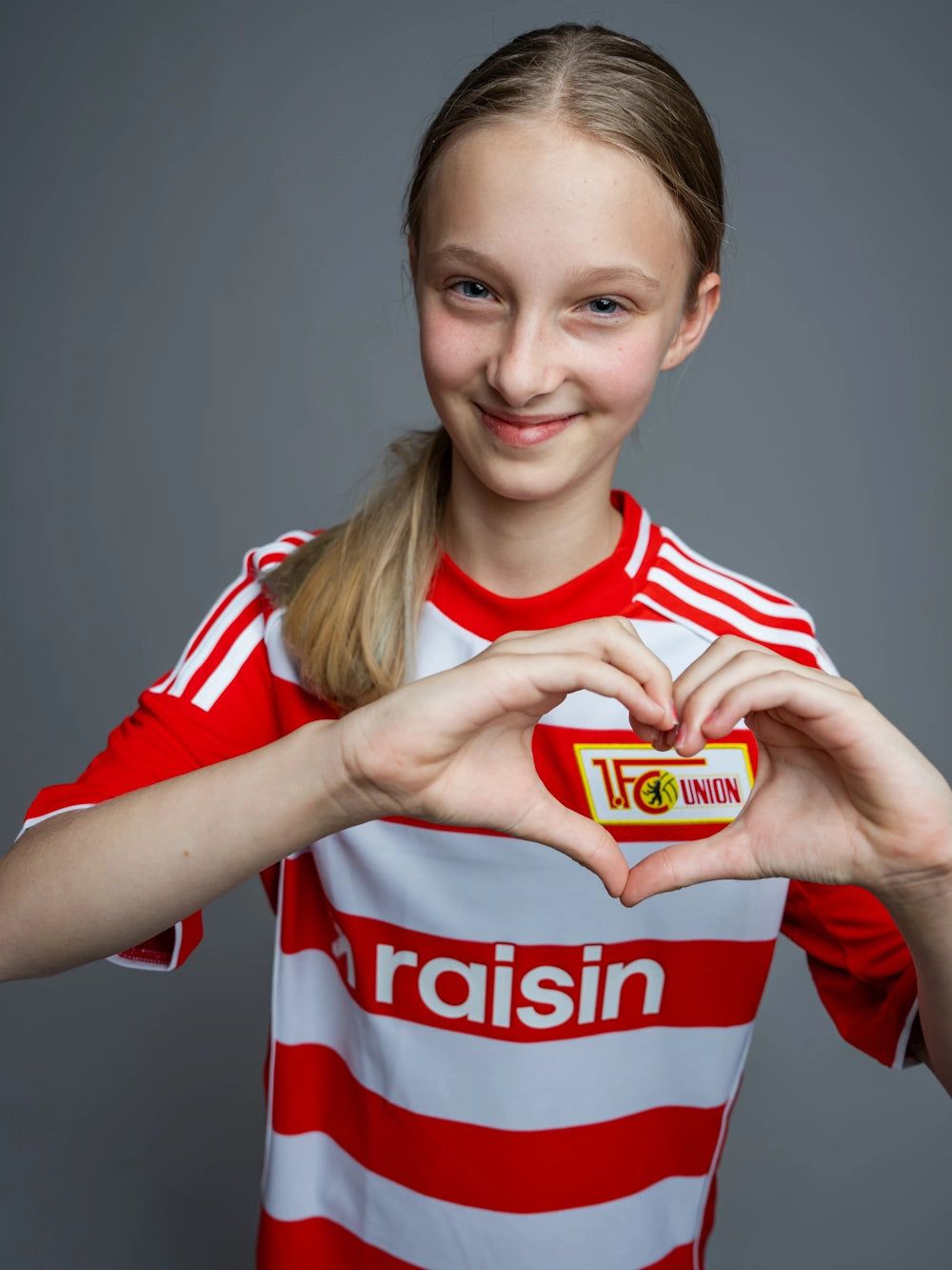 Ein Mädchen in einem rot-weißen Fußballtrikot hält beide Hände in Herzform mit dem Emblem des Vereins TSG Union.