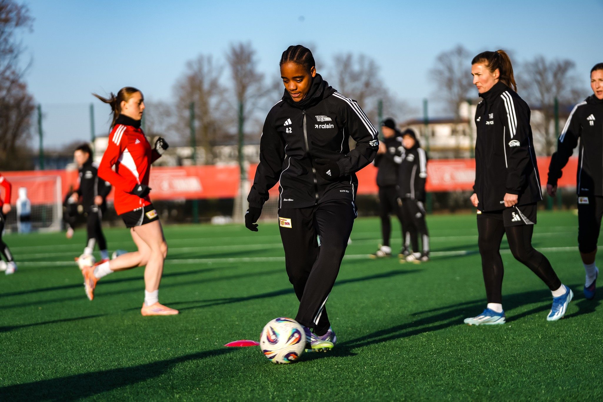 Fußballtraining auf einem Rasenplatz, mehrere Spielerinnen in Trainingskleidung, eine Spielerin dribbelt den Ball.