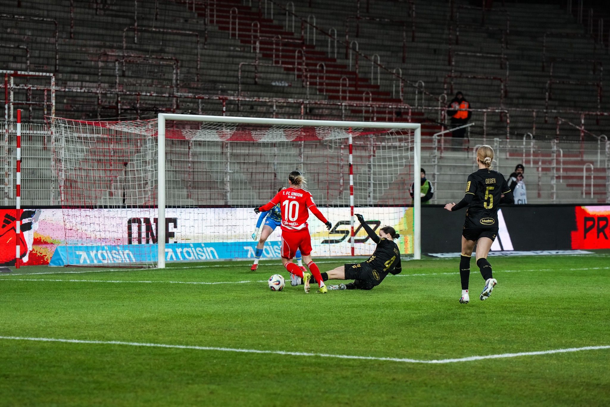 Eine Fußballspielszene, in der ein Spieler mit der Nummer 10 vor dem Torwart ausweicht, während ein weiterer Spieler zuschaut.