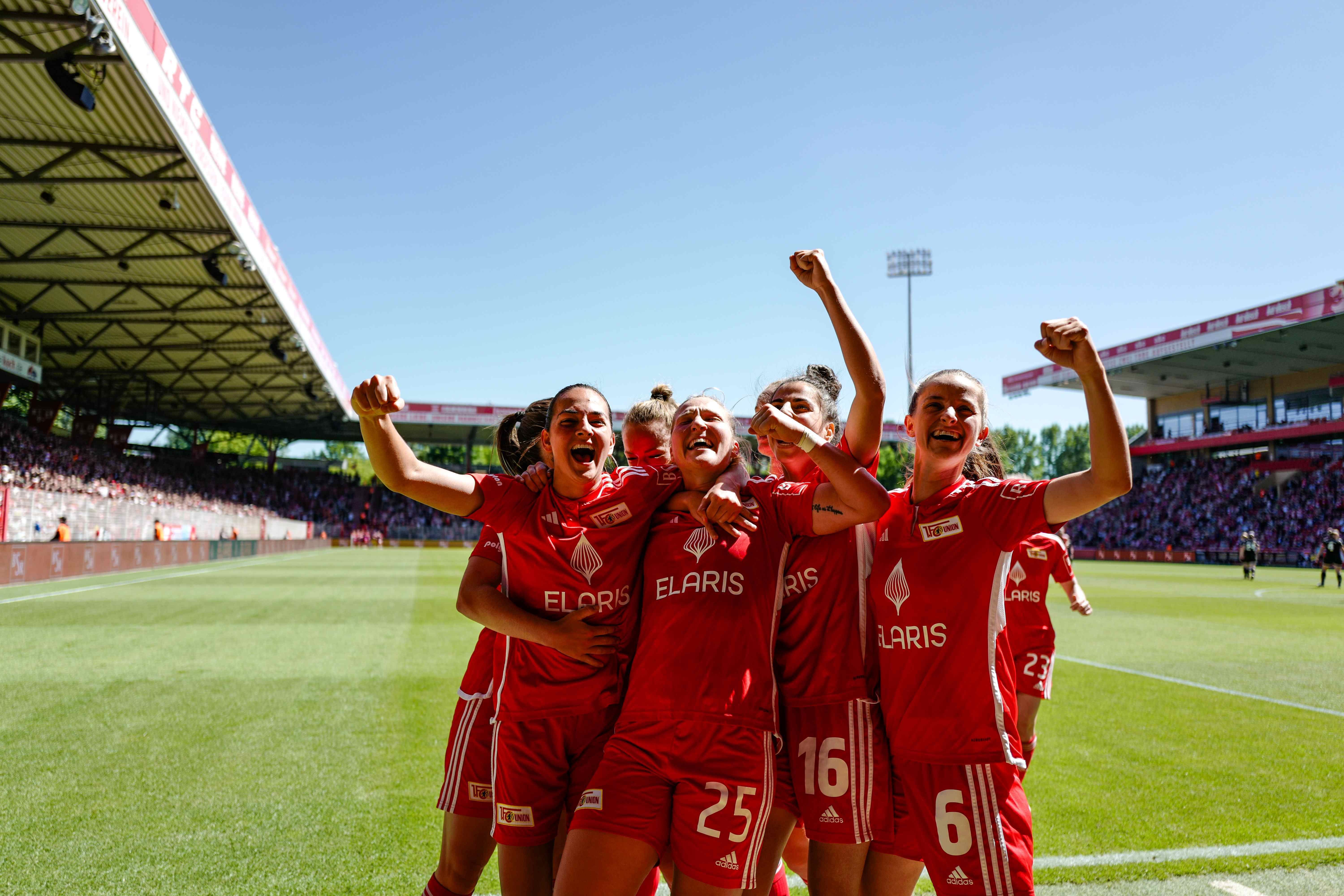 Fünf jubelnde Fußballspielerinnen in roten Trikots feiern einen Torerfolg auf dem Spielfeld. Sonniger Tag im Stadion.