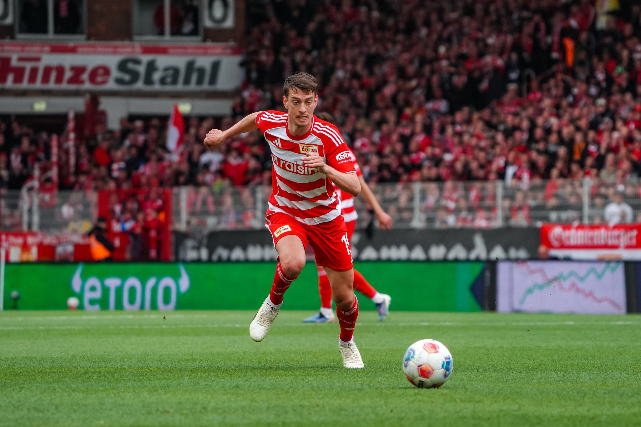 Spieler in rot-gestreifter Fußballuniform sprintet über das Feld, um den Ball in einem Stadion mit jubelndem Publikum.