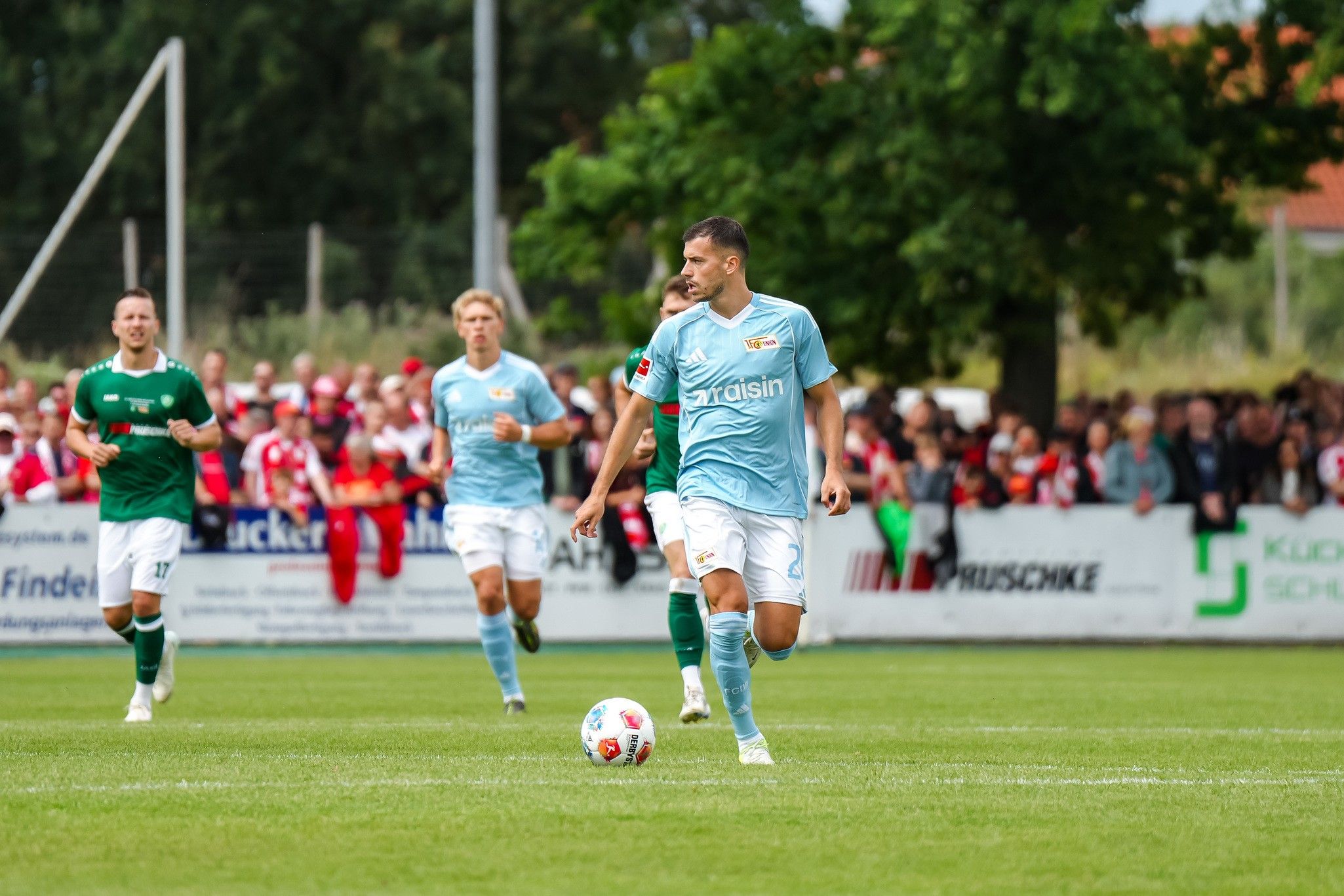 Fußballer in hellblauem Trikot laufen auf einem Spielfeld mit Zuschauern im Hintergrund.