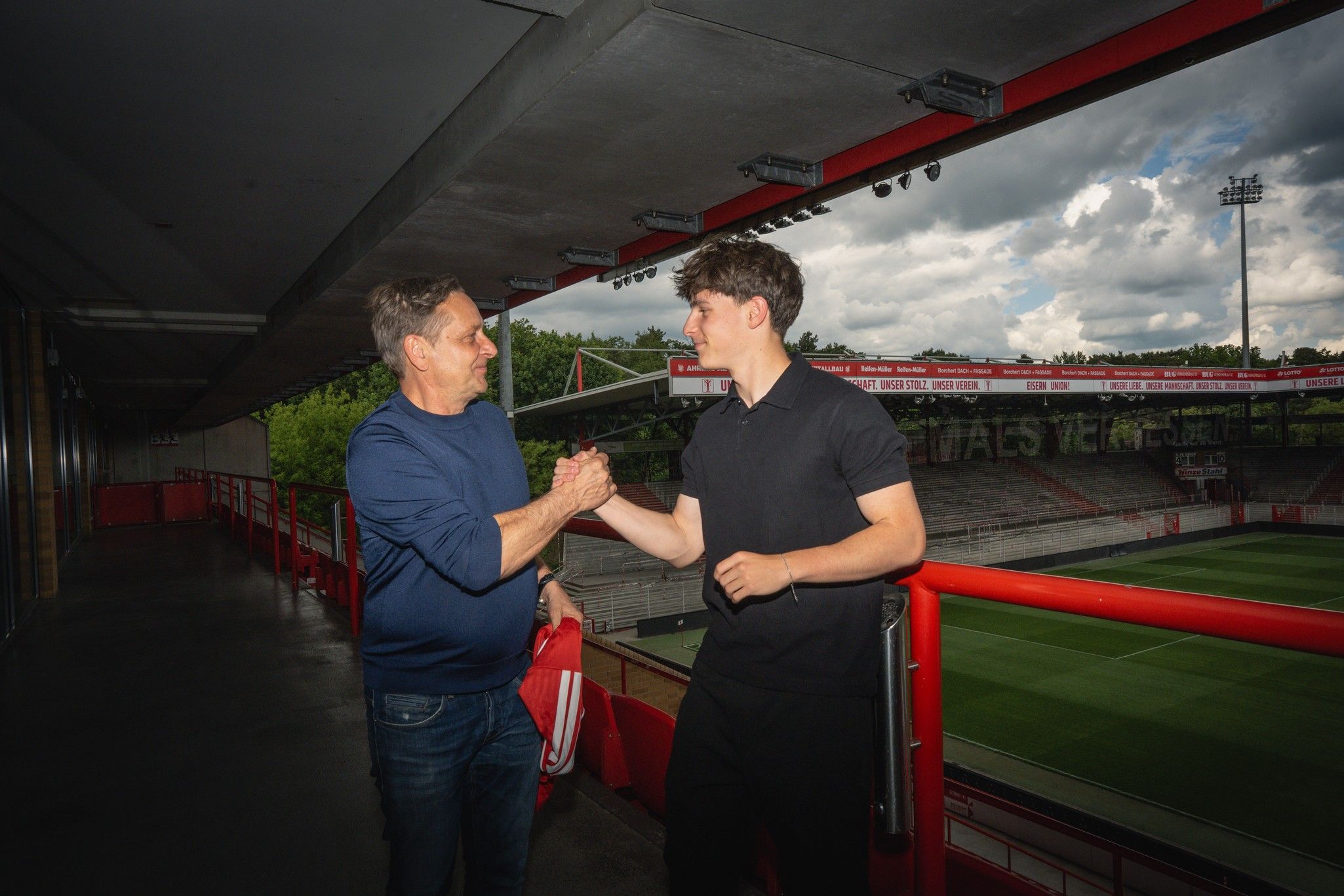 Ein Mann und ein junger Mann schütteln sich die Hände auf einer Stadiontribüne mit Rasen im Hintergrund.