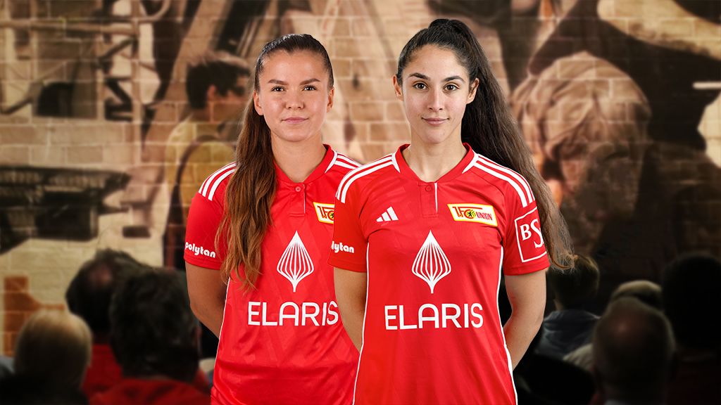Zwei Fußballspielerinnen in roten Trikots mit dem Logo "ELARIS" stehen vor einem Publikum.