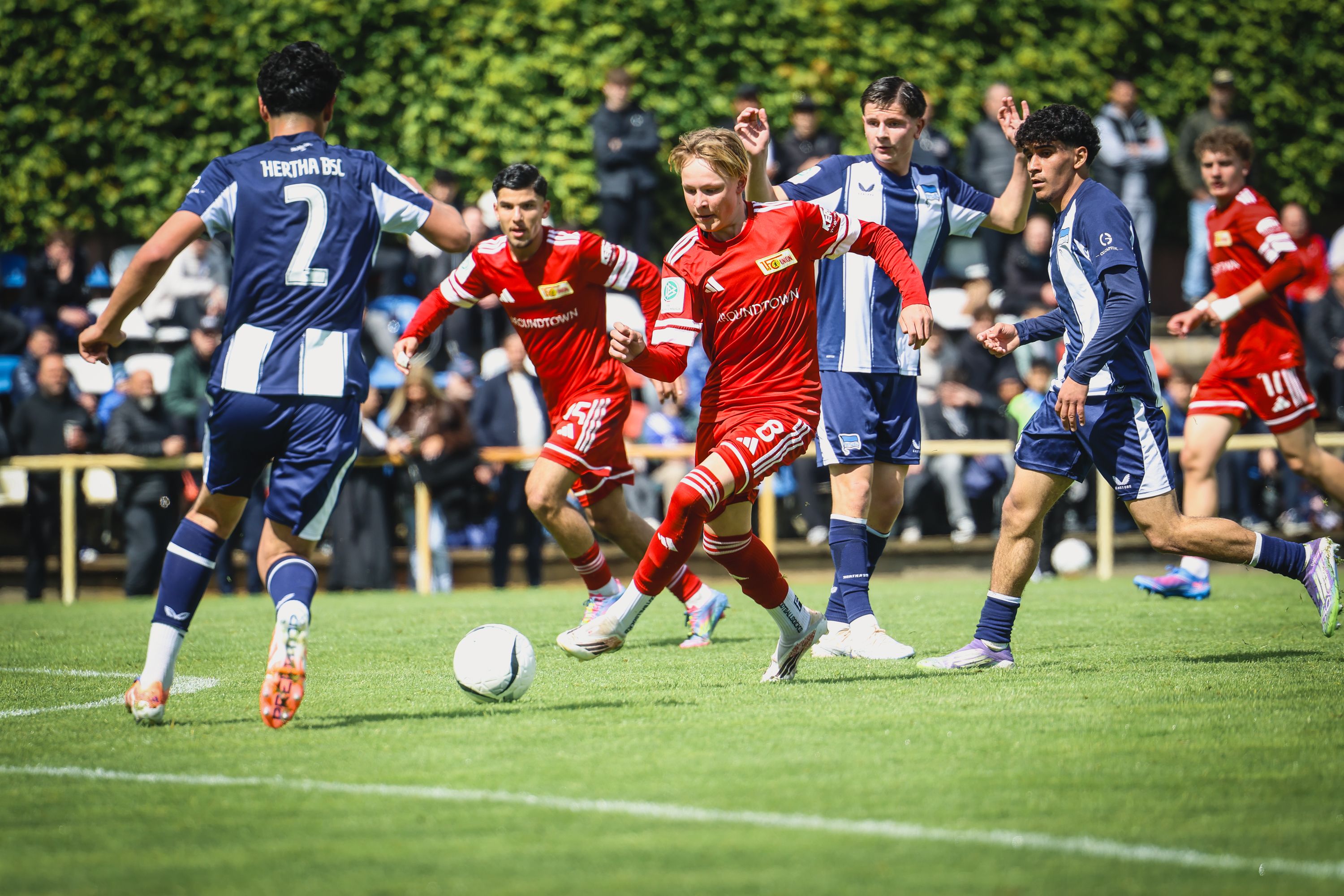 Spielszene eines Fußballmatches mit Spielern in roten und blauen Trikots auf einem Rasenfeld. Zuschauer im Hintergrund.