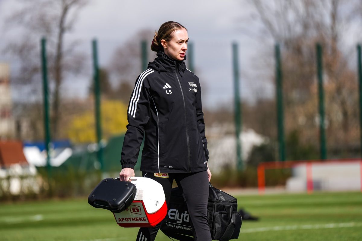 Frau in sportlicher Kleidung trägt eine medizinische Tasche und steht auf einem Fußballfeld.