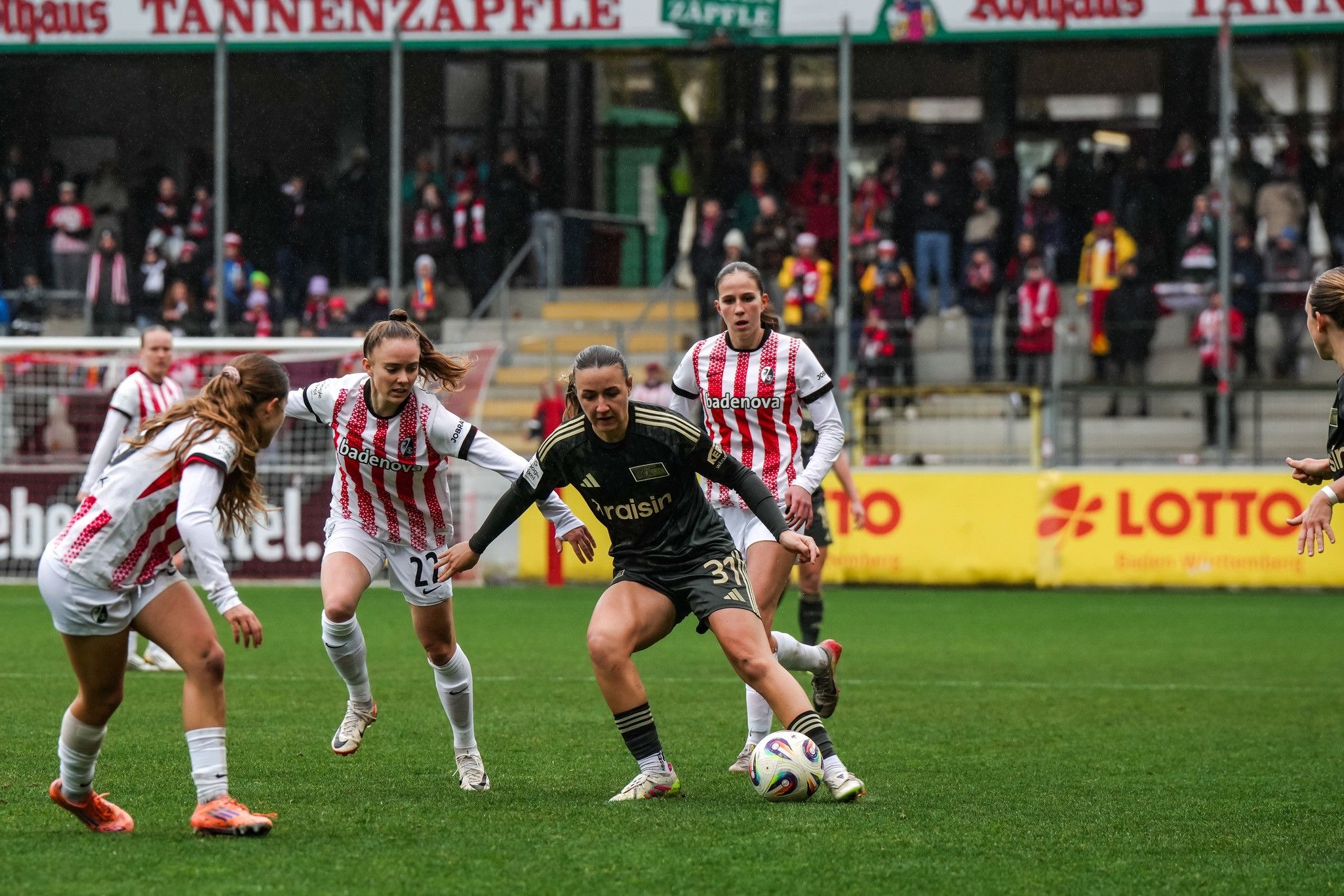 Eine Torhüterin im Schwarz dribbelt den Ball, während zwei gegnerische Spielerinnen ihn verfolgen. Zuschauer im Hintergrund.