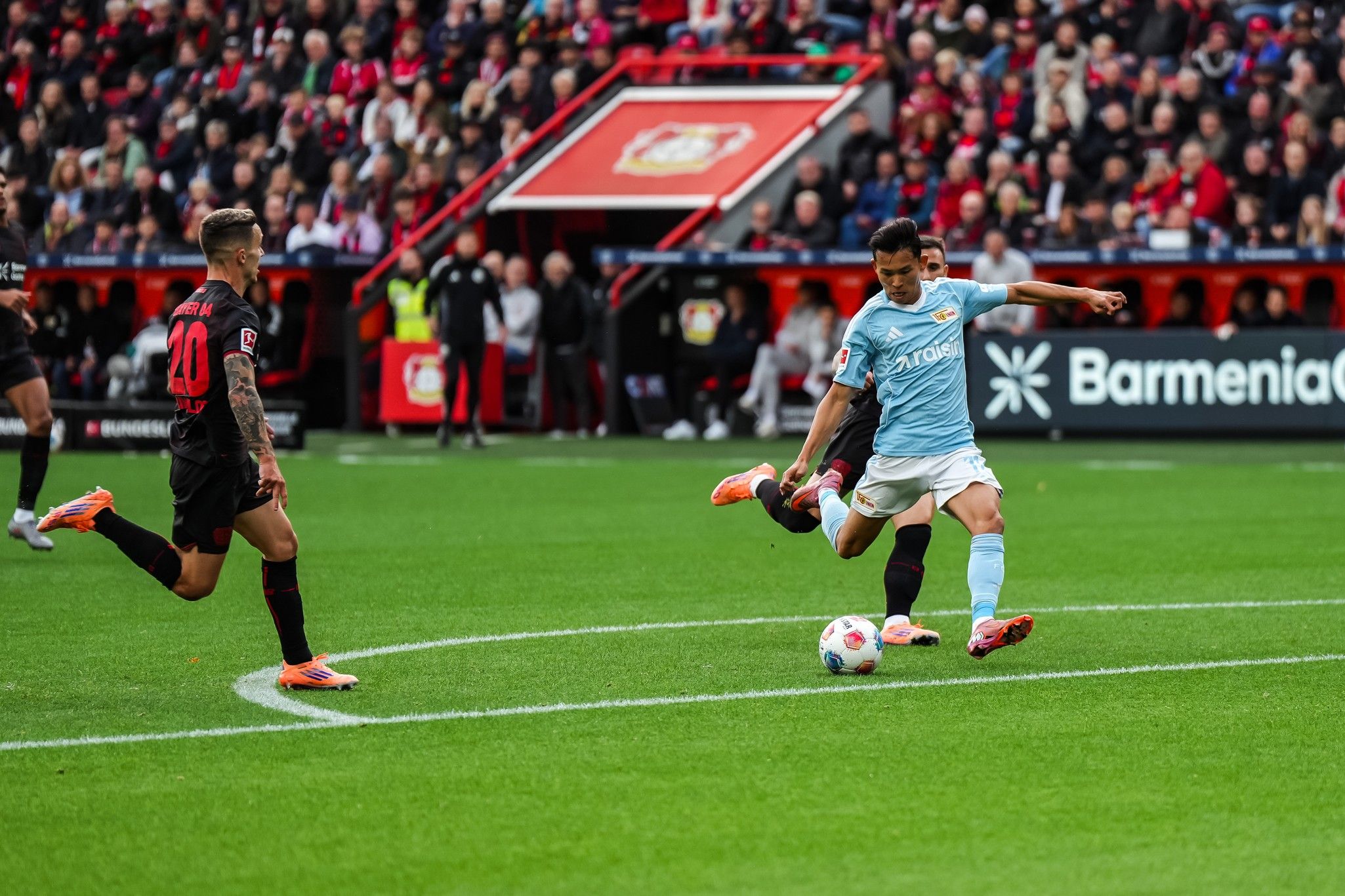 Spielszene im Fußball: Spieler in hellblauer Kleidung schießt den Ball, während ein Gegner in schwarz daneben steht. Zuschauer im Hintergrund.