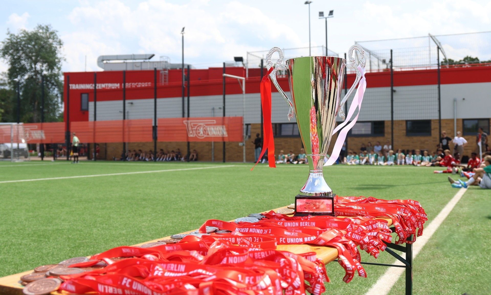 Ein silberner Pokal steht auf einem Tisch mit roten Medaillen auf einem Fußballplatz bei schönem Wetter.