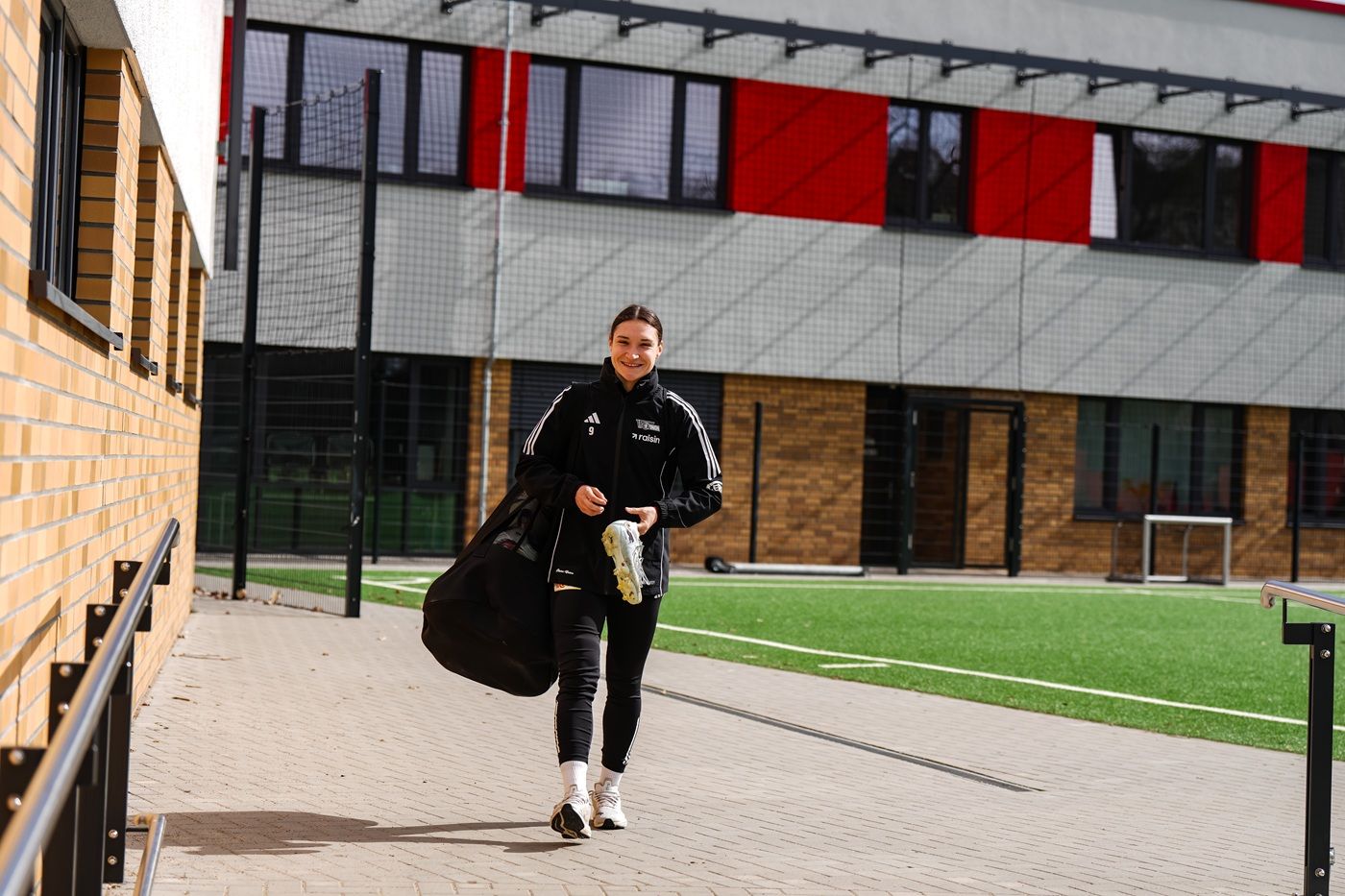 Eine sportliche Frau in Trainingskleidung geht mit einer Tasche und Getränk auf einem Sportgelände.