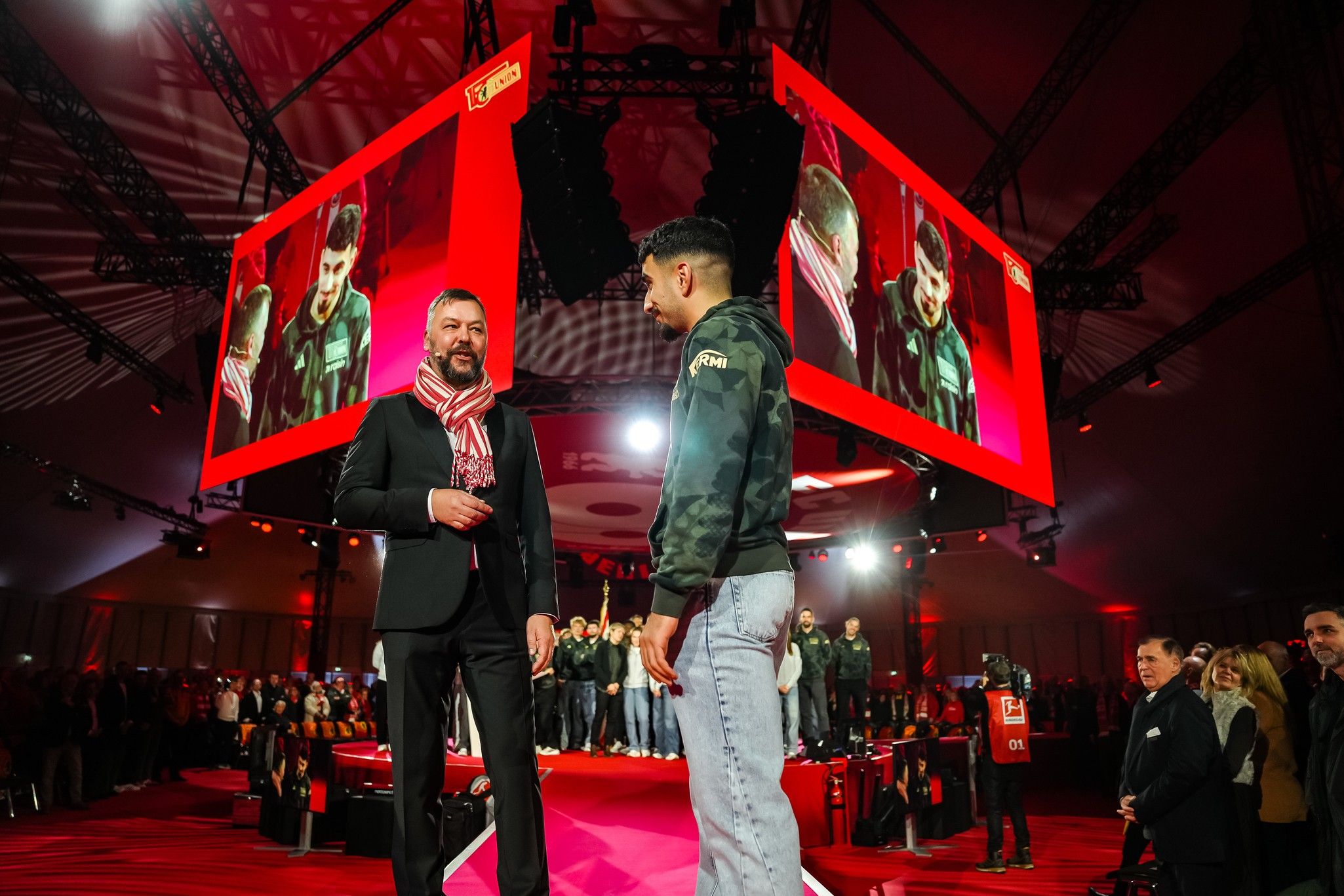 Eine Veranstaltung mit einer Bühne und großen Bildschirmen, auf denen ein Gespräch zwischen zwei Personen zu sehen ist. Publikum im Hintergrund.