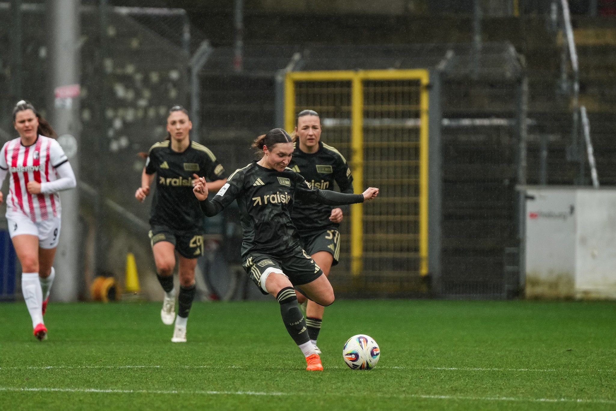 Eine Spielerin in schwarzem Trikot schießt den Ball, während ihre Mitspielerinnen im Hintergrund stehen. Rasen und Torwand sichtbar.