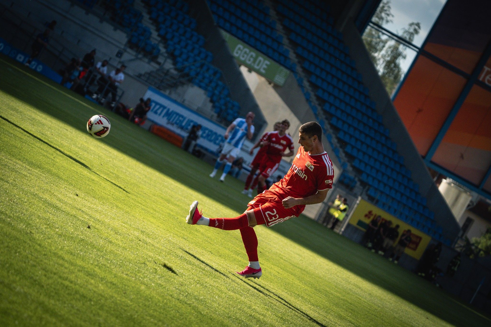 Ein Fußballspieler in roter Ausrüstung schlägt einen Ball auf einem Stadionrasen. Im Hintergrund sind einige Zuschauer und Spieler zu sehen.