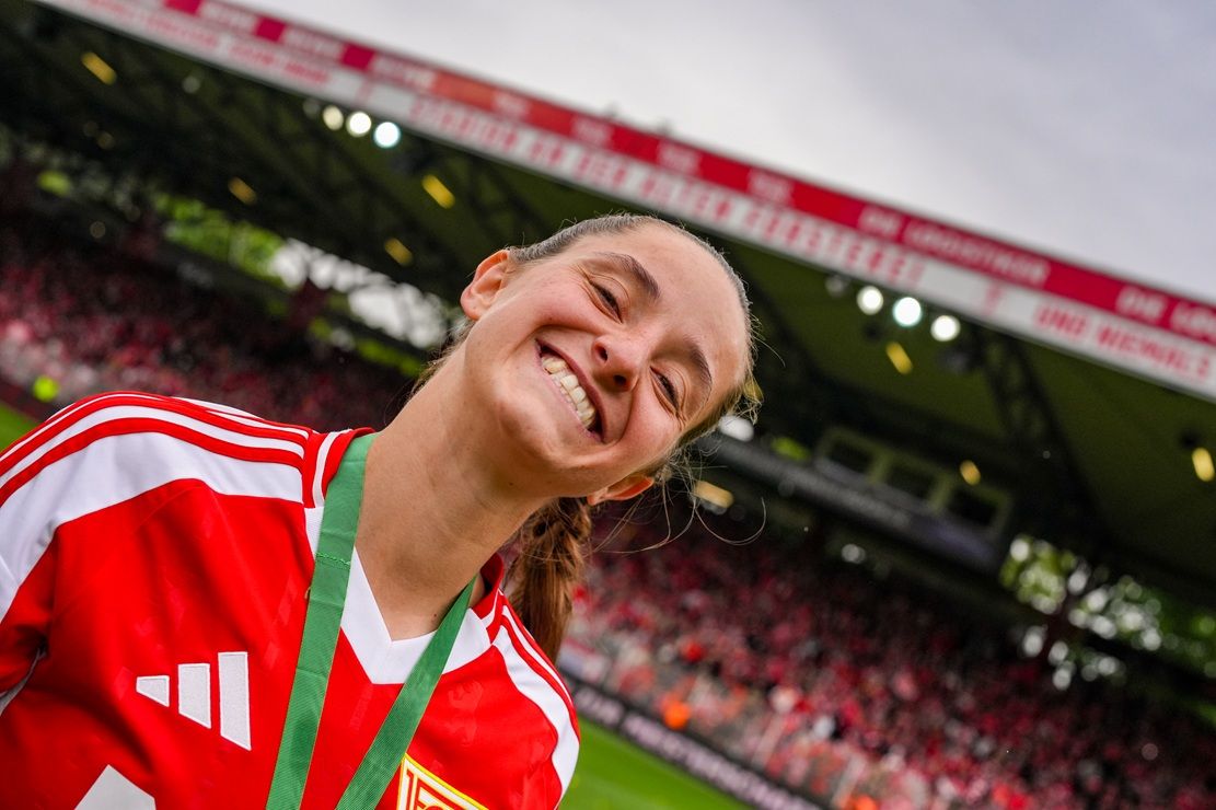 Eine lächelnde Sportlerin in einem roten Trikot steht im Stadion, im Hintergrund ein Publikum und eine Tribüne.