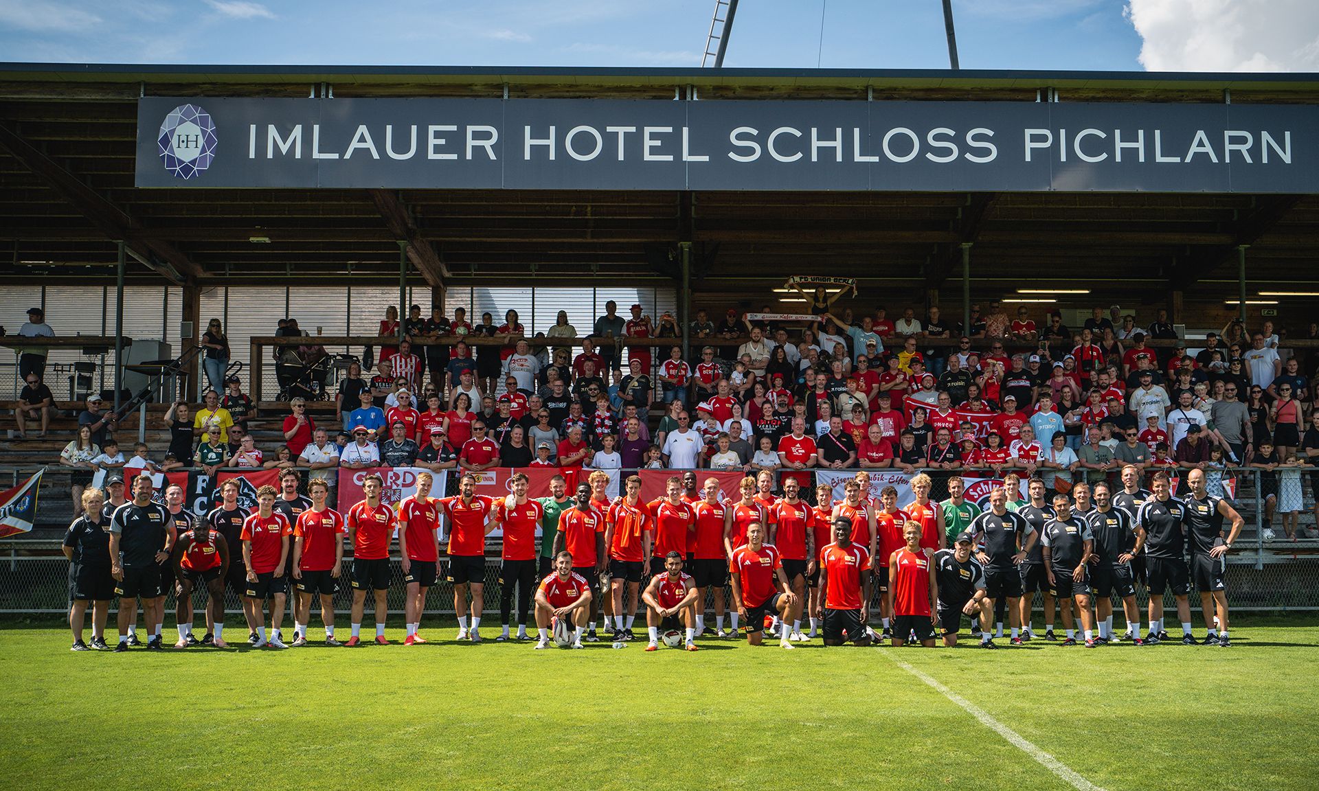 Gruppenshot von Sportlern in roten und schwarzen Trikots auf einem Fußballfeld, mit Publikum im Hintergrund.
