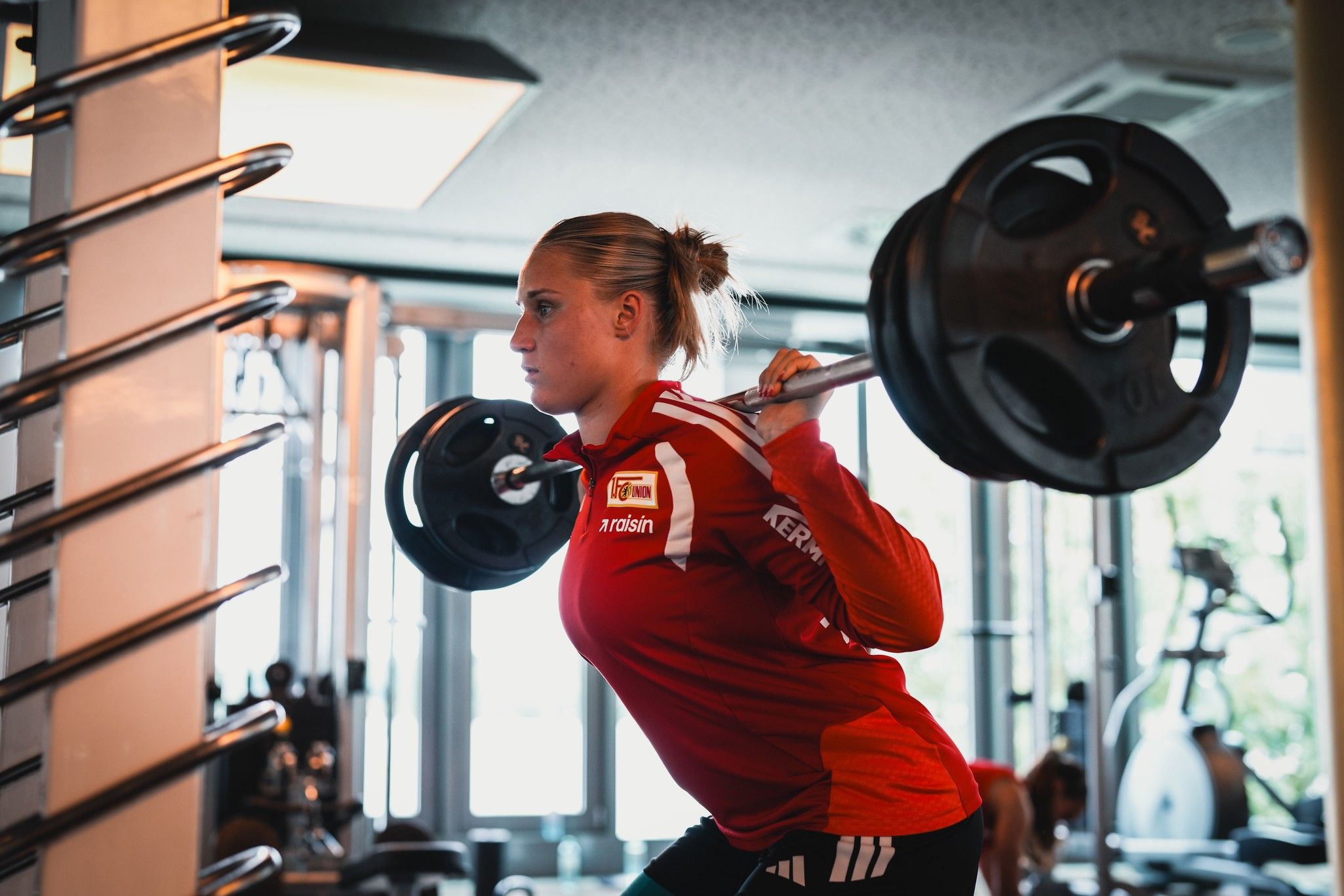 Eine sportliche Person in roter Trainingskleidung hebt eine Langhantel im Fitnessstudio.