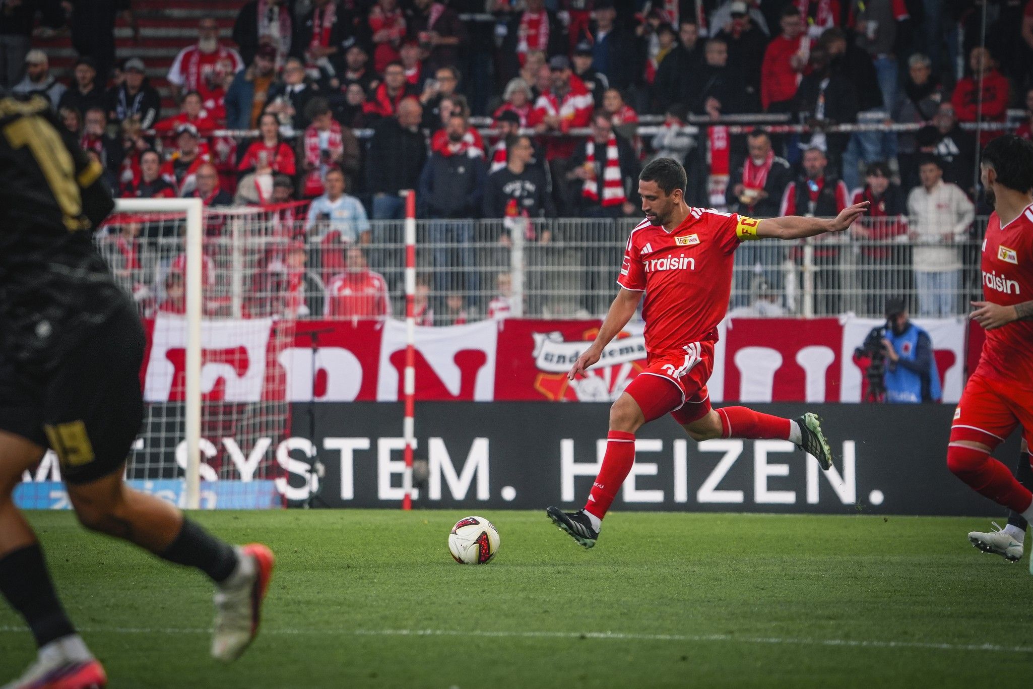 Spieler in rotem Trikot führt einen Schuss auf das Tor aus, während Fans im Hintergrund jubeln.