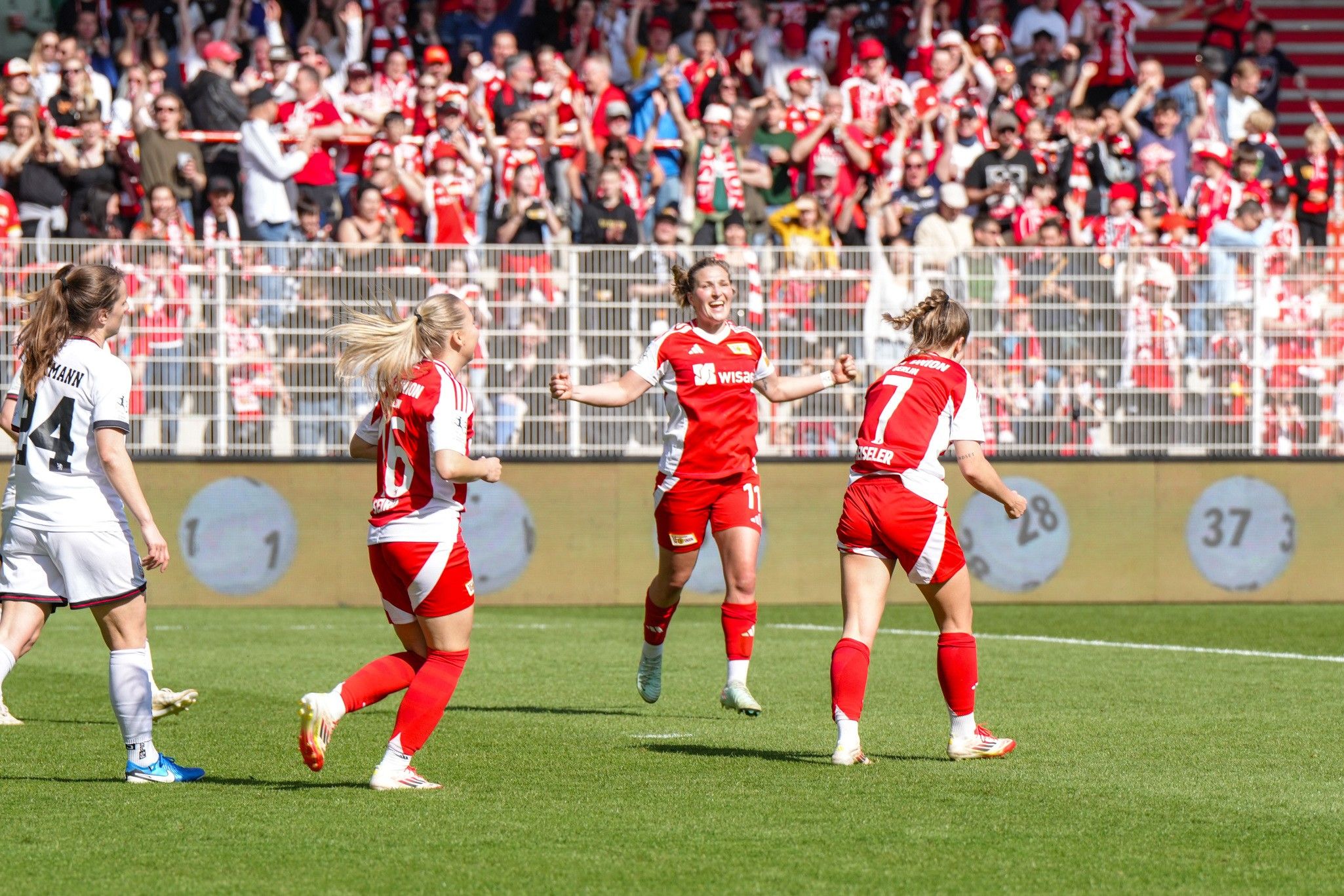 Spielerinnen in roten Trikots feiern ein Tor, während Zuschauer im Hintergrund jubeln. Es ist ein lebhaftes Fußballspiel.