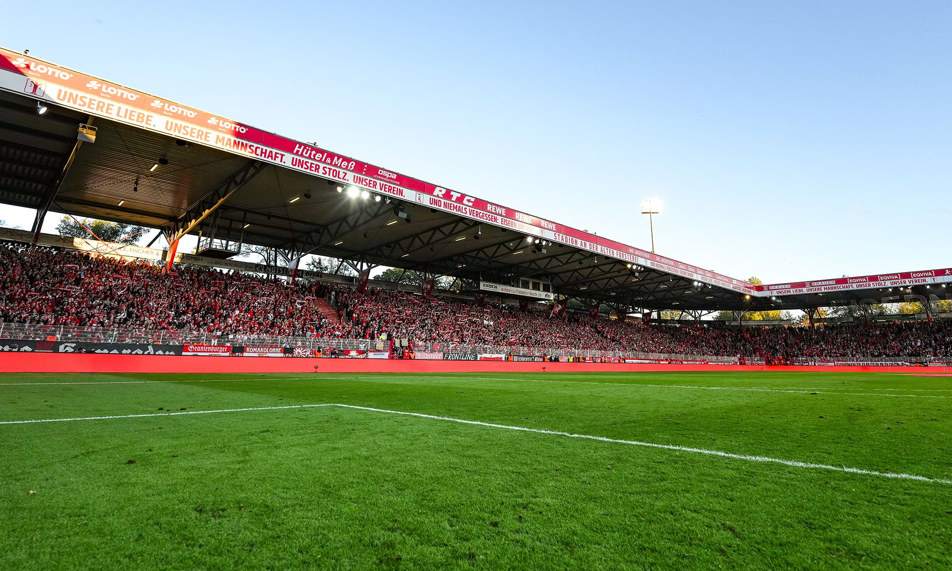 Blick auf ein vollbesetztes Fußballstadion mit grüner Spielfläche und Tribünen unter blauem Himmel.