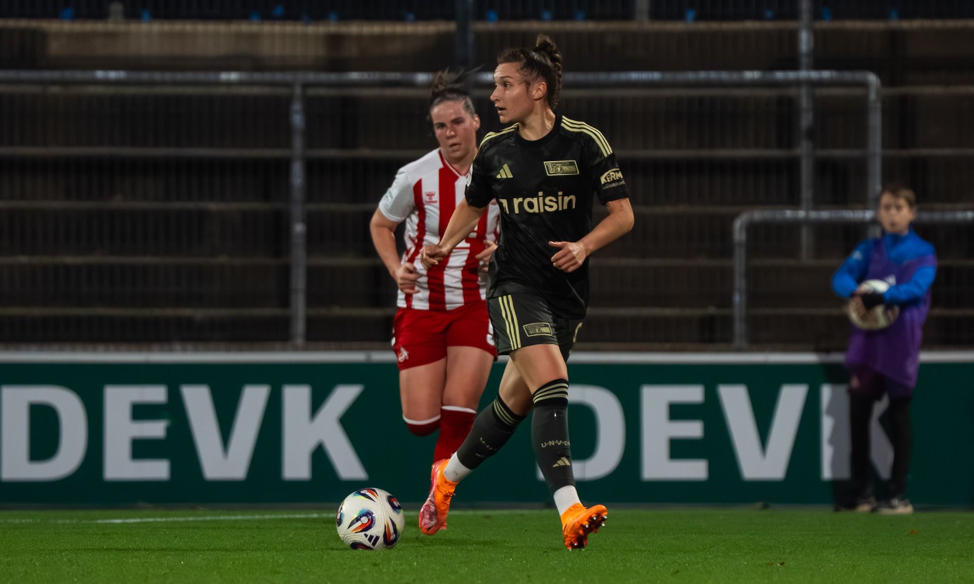 Eine Fußballspielerin in schwarzem Trikot mit goldenen Details dribbelt den Ball, während eine Gegnerin in rot-weißem Trikot verfolgt.