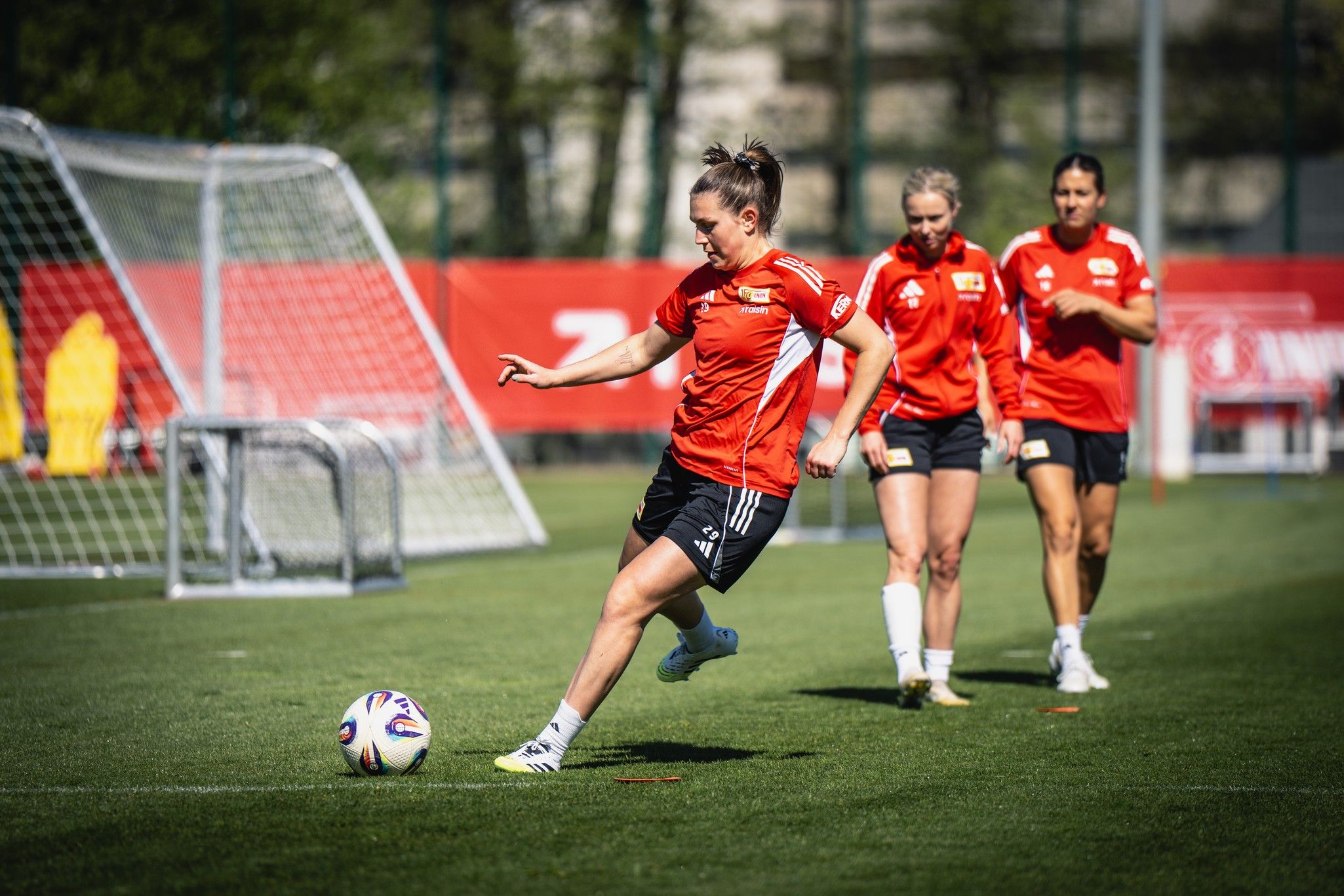 Eine Fußballspielerin in roter Trainingskleidung schießt einen Ball, während weitere Spielerinnen im Hintergrund stehen.