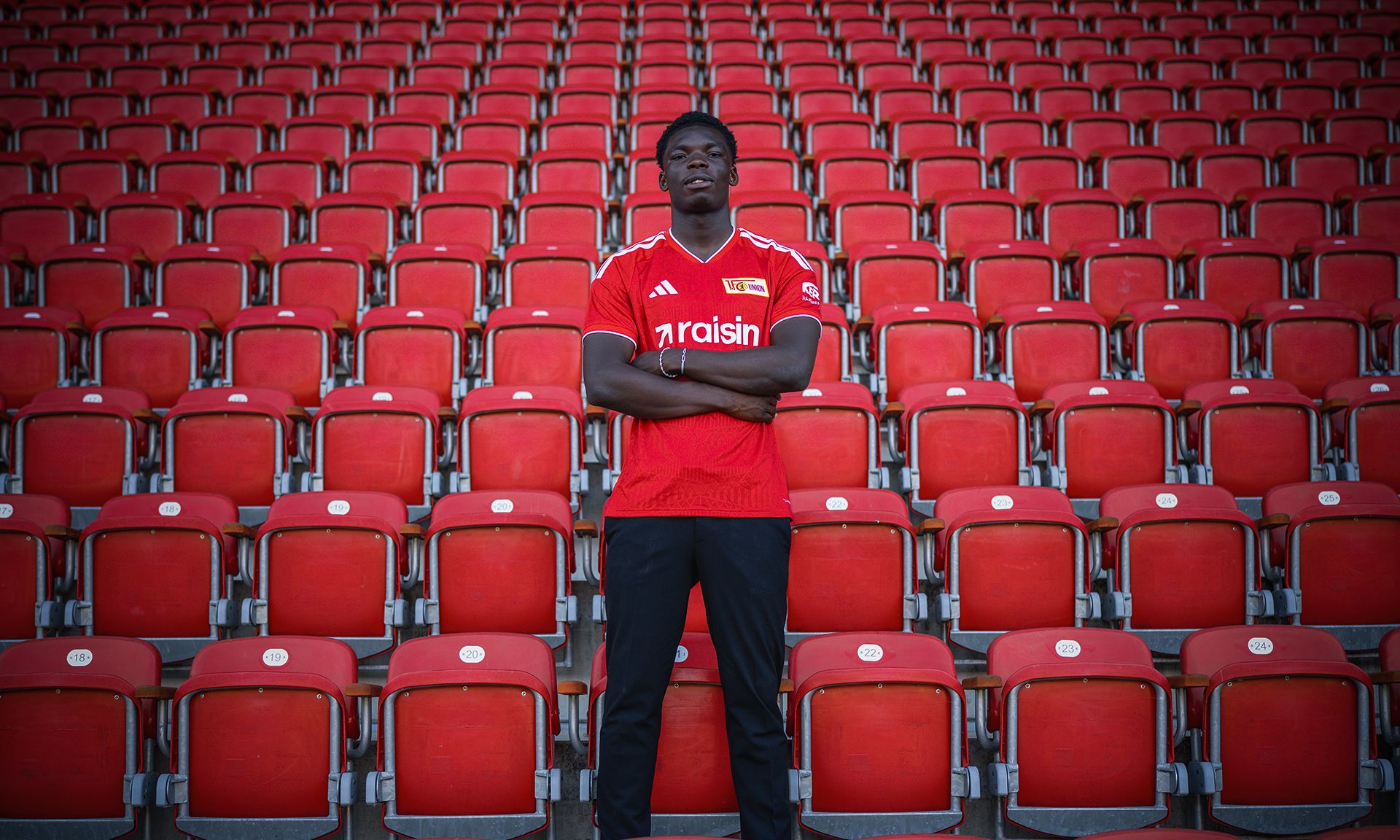 Eine Person in rotem Trikot steht aufrecht vor einer leeren Reihe roter Stadionplätze.