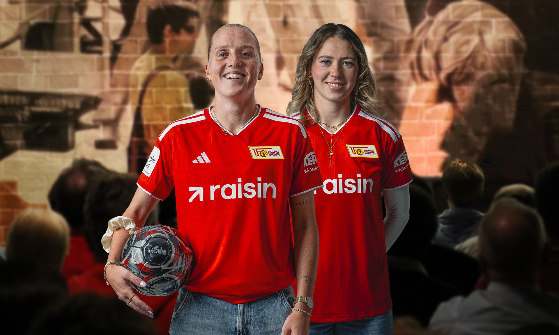 Zwei Personen in roten Trikots mit dem Aufdruck "raisin", stehen lächelnd mit einem Fußball in der Hand vor einem Publikum.