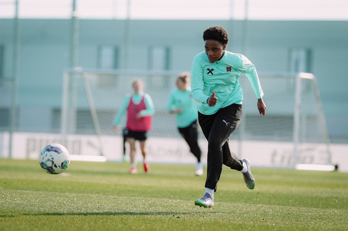 Eine junge Sportlerin in mintgrüner Trainingskleidung sprintet mit einem Fußball auf einem grünen Sportplatz.