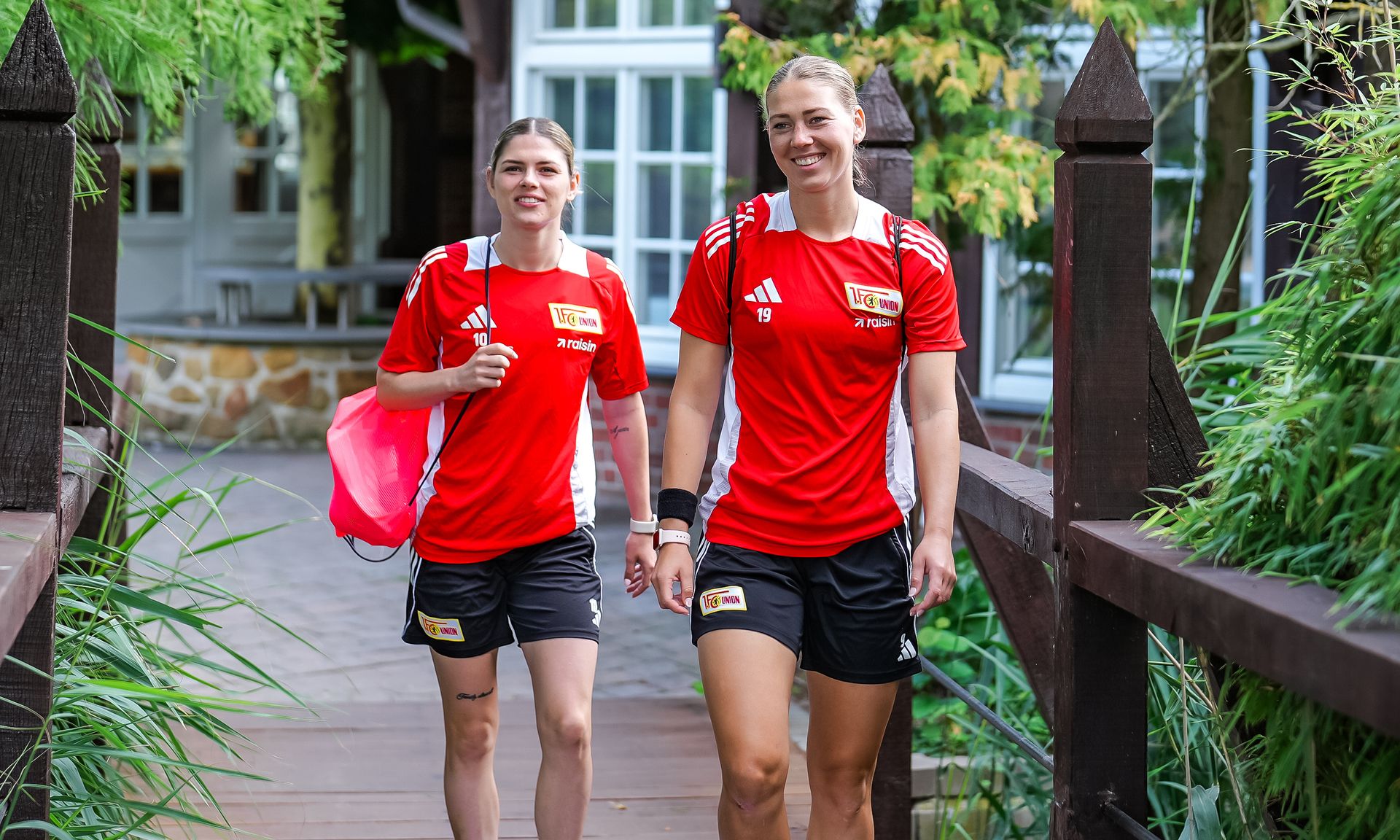 Zwei sportlich gekleidete Frauen in roten Shirts gehen lächelnd auf einem Brückengang entlang, umgeben von Pflanzen.