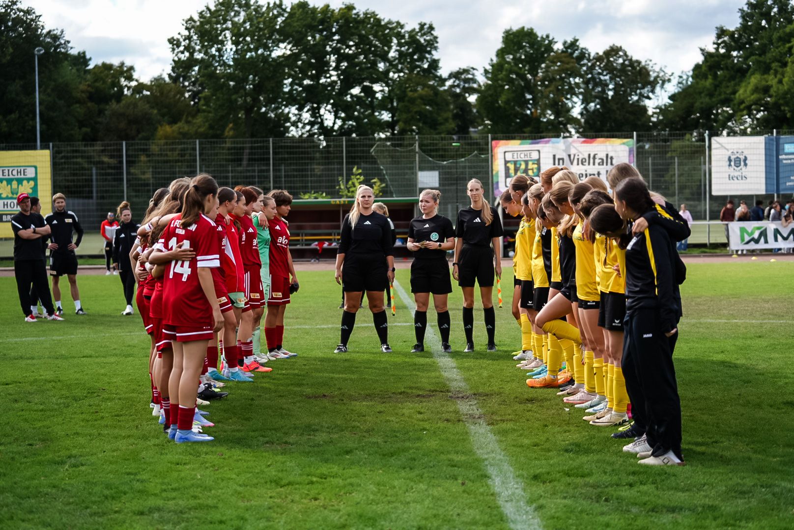 Mädchenfußballmannschaften stehen in zwei Reihen auf dem Spielfeld, bereit für ein Spiel. Schiedsrichterin steht in der Mitte.