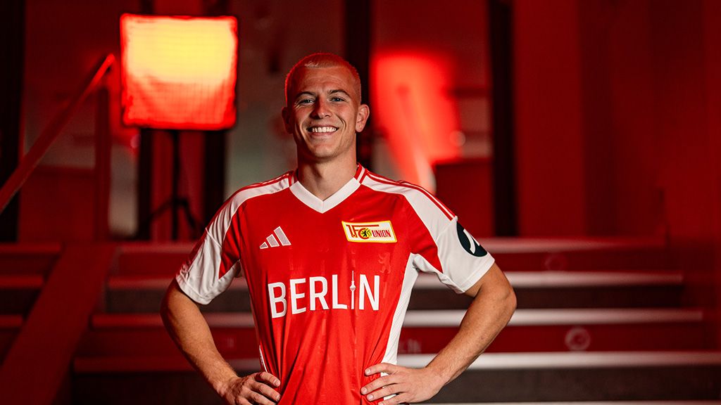 Ein lächelnder Mann in einem roten Fußballtrikot mit der Aufschrift "BERLIN" steht vor einem roten Hintergrund.
