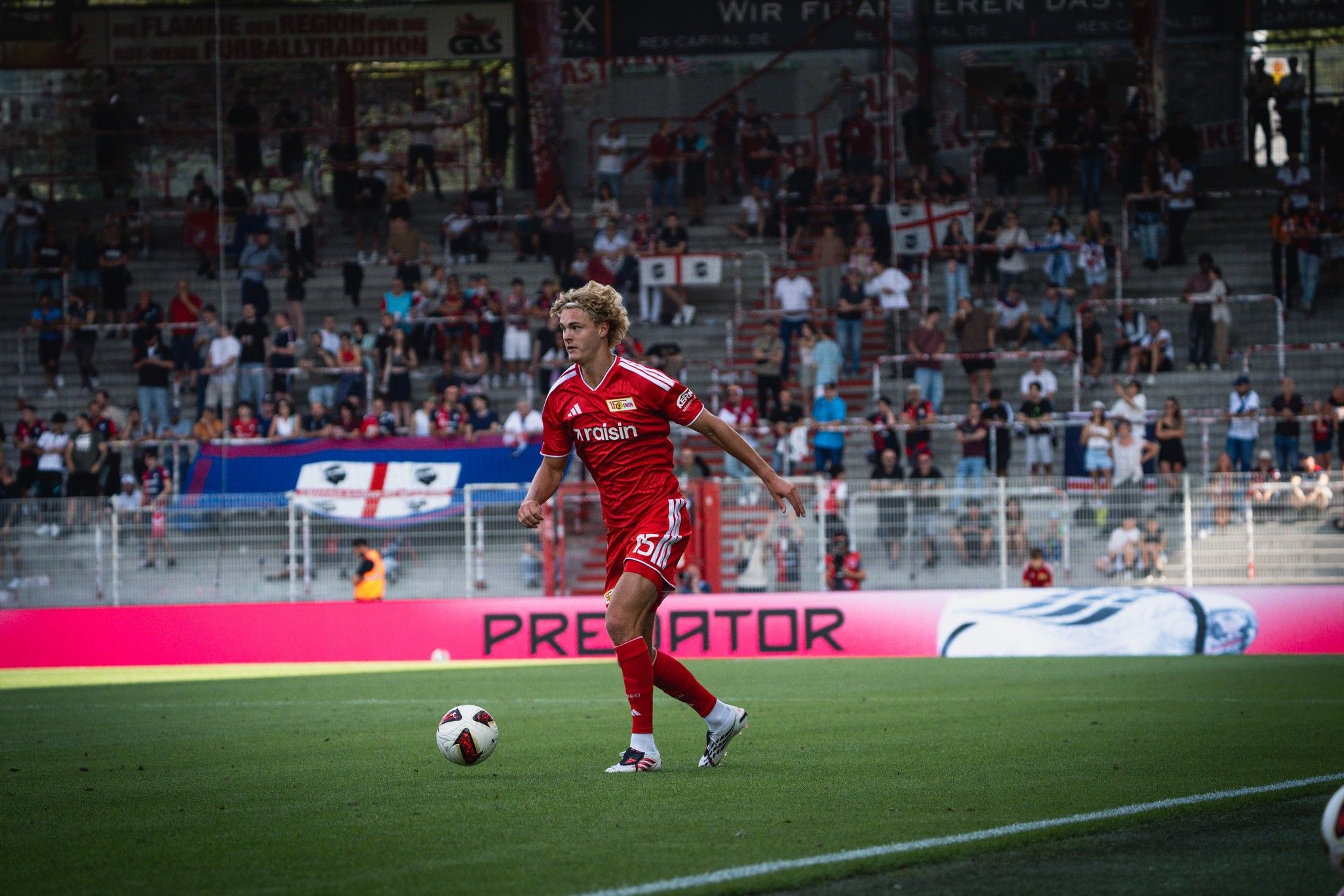 Ein Fußballer in roter Trikotausrüstung steht auf dem Spielfeld und bereitet sich auf einen Pass vor, im Hintergrund Fans und Stadion.
