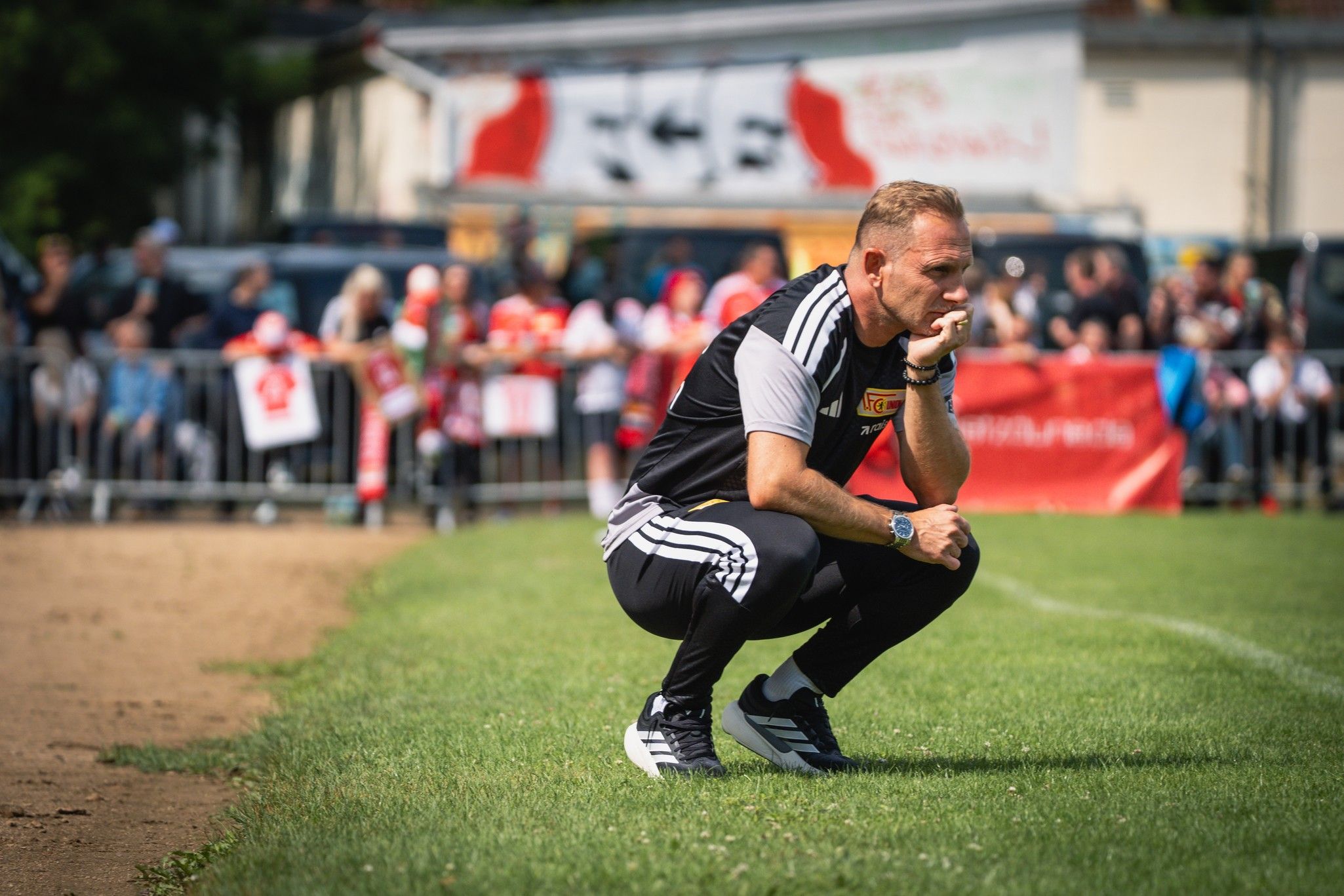 Ein Trainer kniet am Spielfeldrand und beobachtet konzentriert das Spiel, während Zuschauer im Hintergrund zuschauen.
