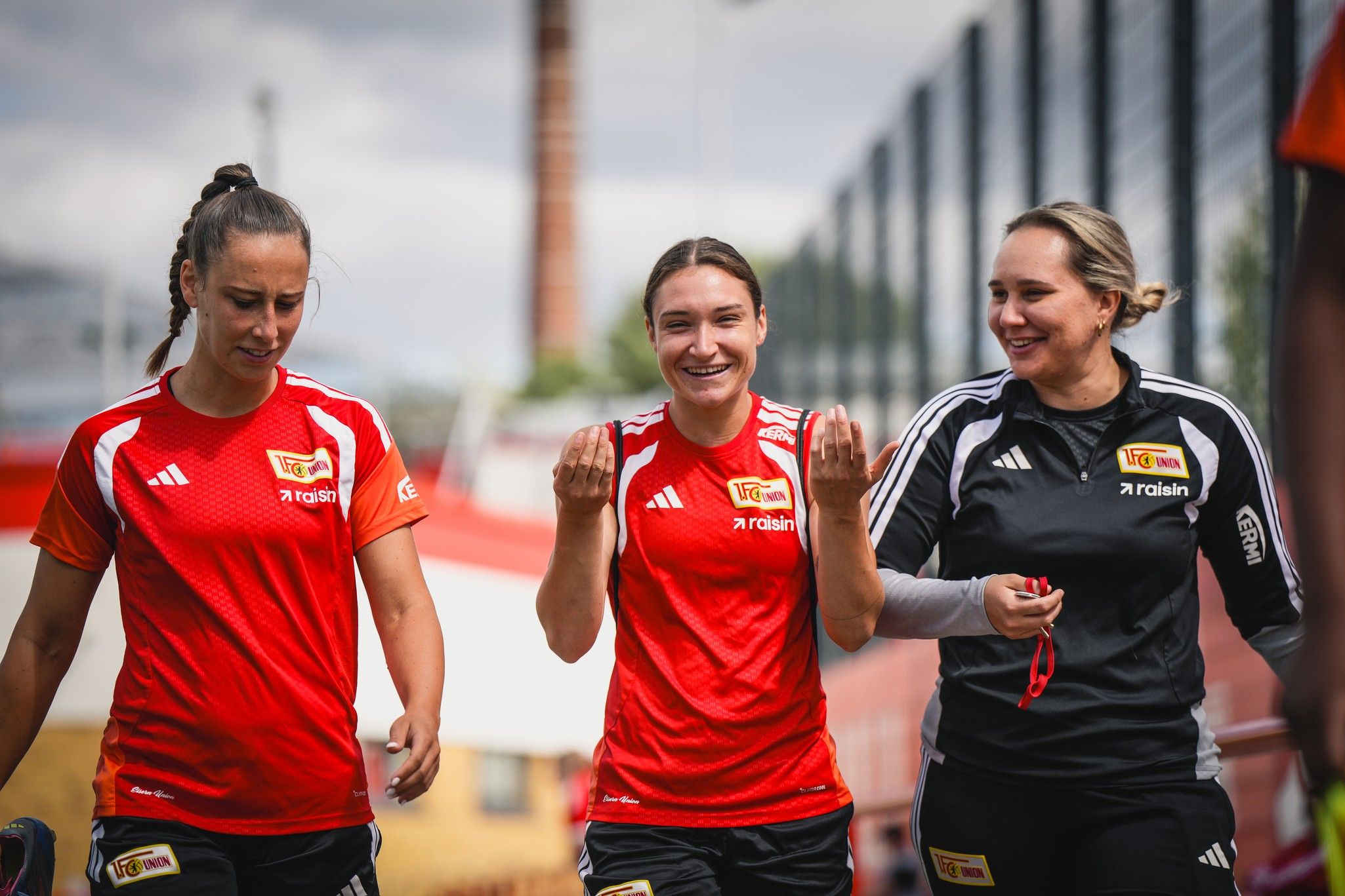 Drei weibliche Fußballspielerinnen in roten Trikots lächeln und gehen gemeinsam auf einem Trainingsgelände.