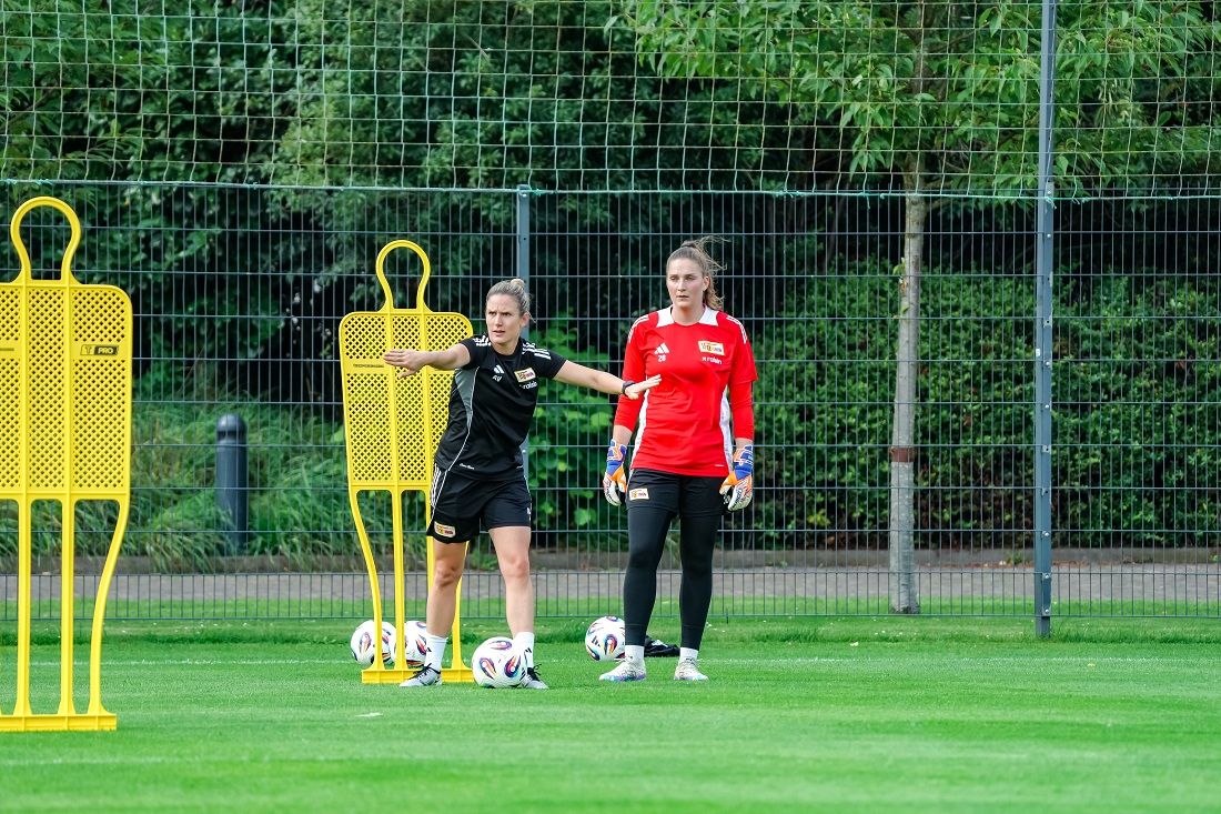 Zwei Fußballspielerinnen stehen auf einem Trainingsplatz, umgeben von Trainingsdummy und Bällen.