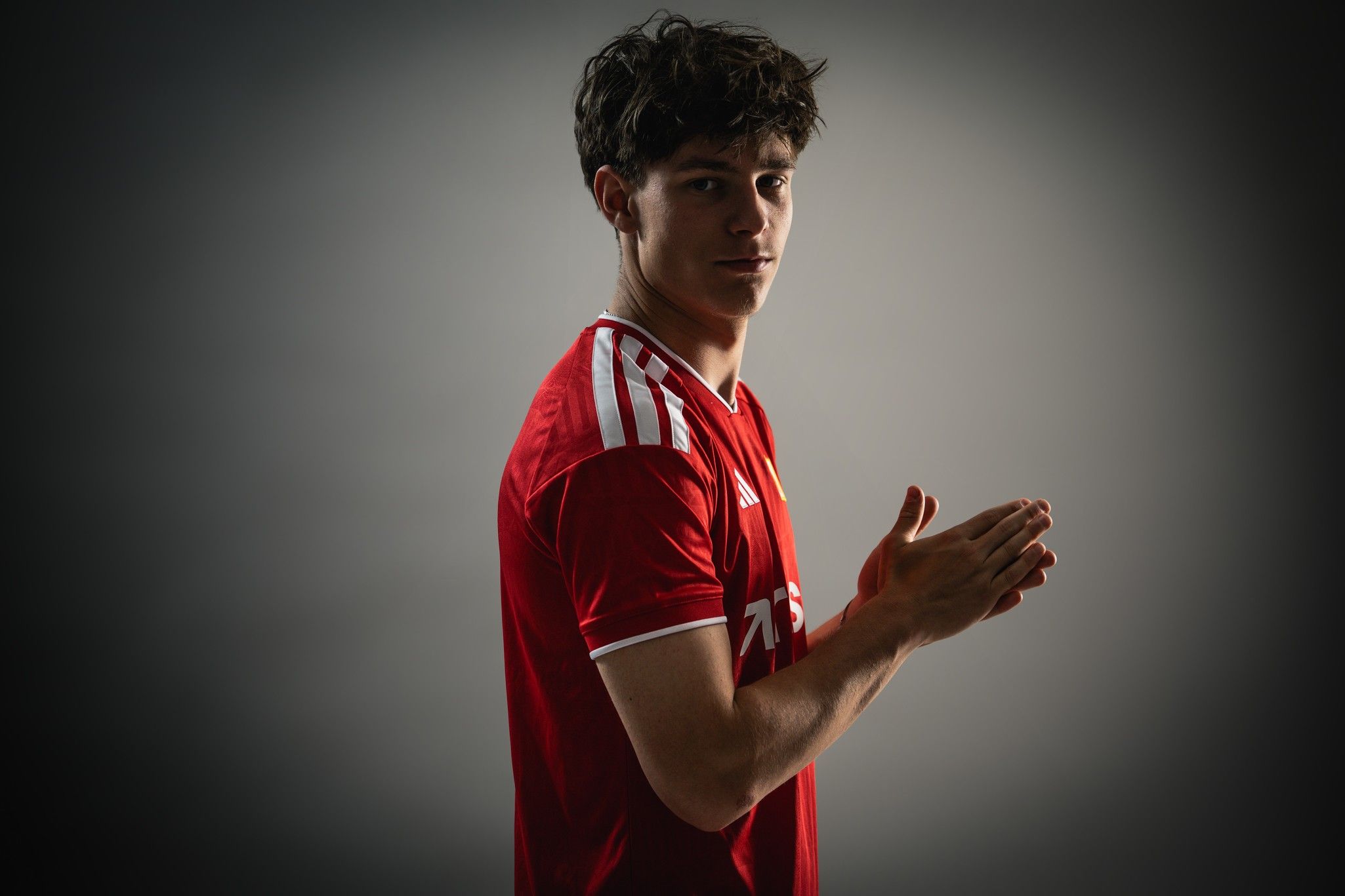 Junger Mann in rotem Fußballtrikot steht seitlich mit leichtem Lächeln und klatscht in die Hände vor einem dunklen Hintergrund.