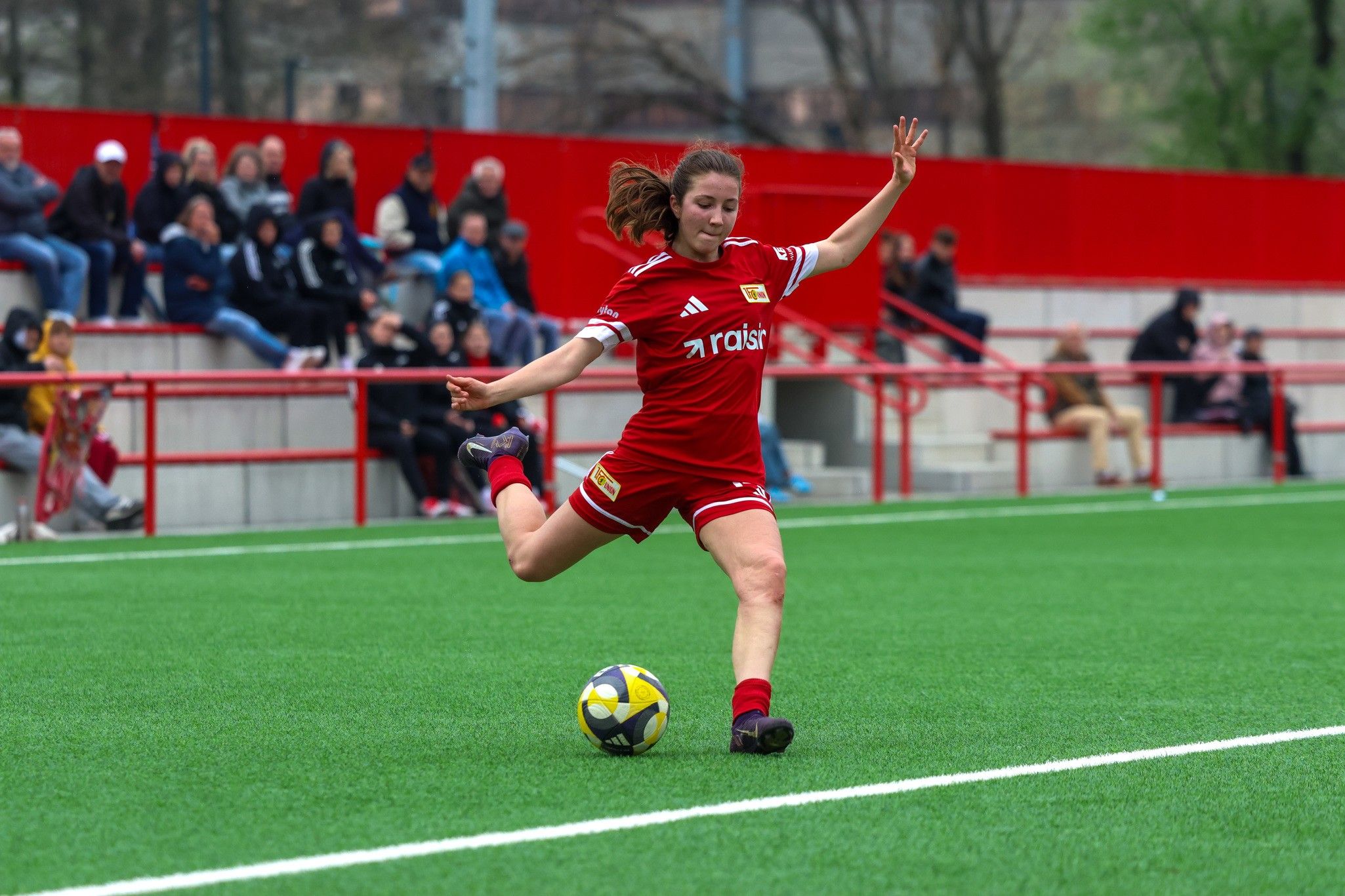 Eine Spielerin in rotem Trikot schießt einen Fußball auf einem grünen Kunstrasenplatz. Zuschauer sitzen auf Tribünen im Hintergrund.