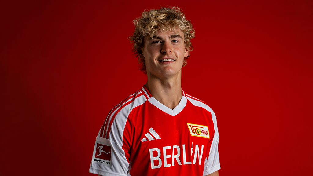 Ein junger Fußballspieler mit Locken trägt ein rotes Trikot mit den Aufdrucken "BERLIN" und "1. FC Union Berlin" vor rotem Hintergrund.