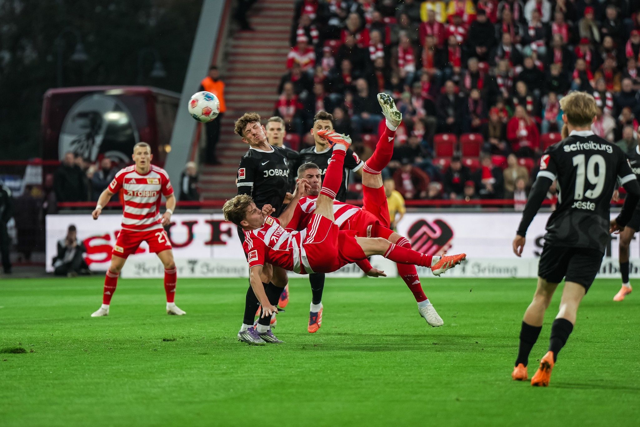 Ein Fußballspieler führt einen akrobatischen Seitfallzieher aus, während zwei andere Spieler im Hintergrund agieren. Fans sind im Stadion sichtbar.