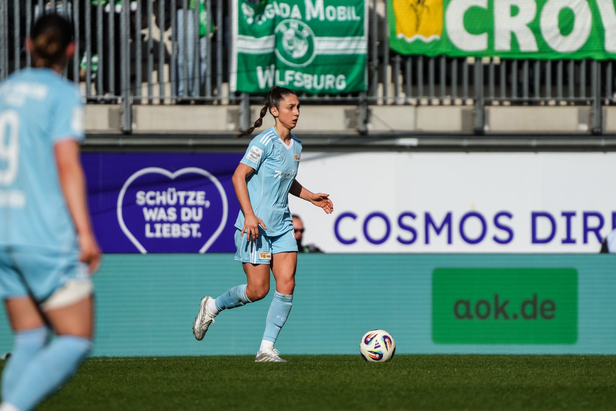 Spielerin in hellblauem Trikot mit Ball auf dem Spielfeld, im Hintergrund Fans und Banner.