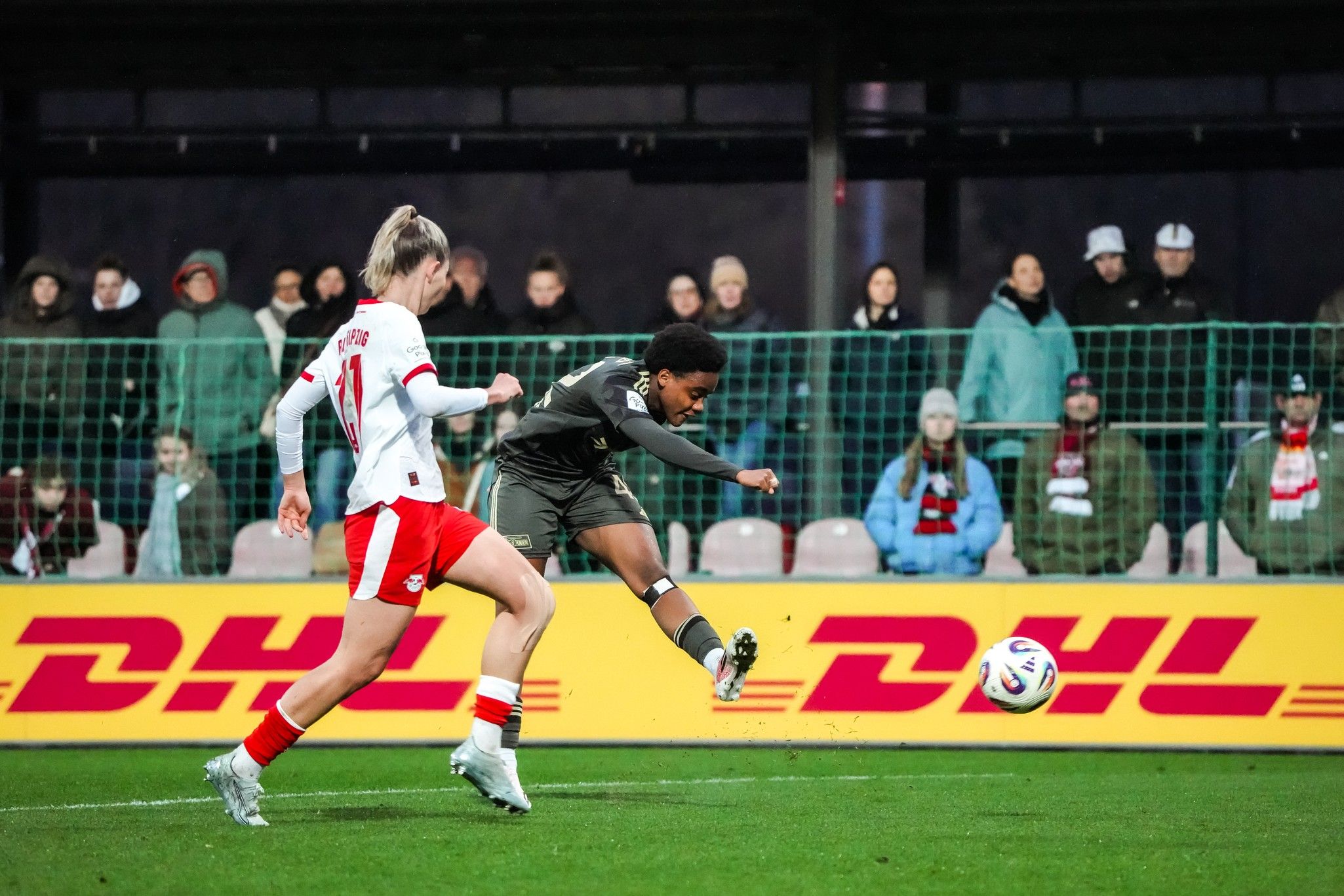 Ein Fußballspiel zeigt eine Spielerin, die den Ball mit einem Schuss Richtung Tor trifft, während eine Gegenspielerin näher kommt.