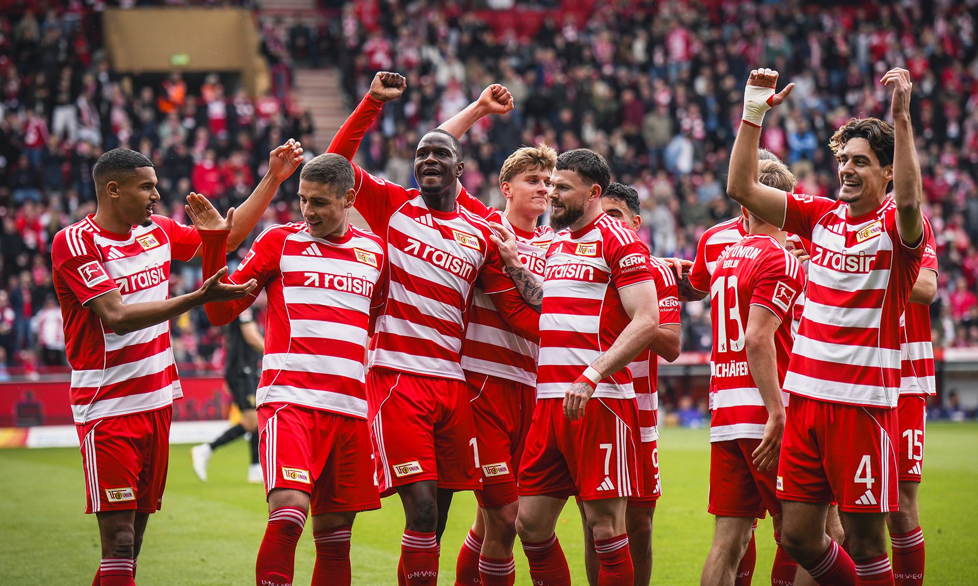 Spieler eines Fußballteams in rot-weiß gestreiften Trikots feiern einen Torerfolg auf dem Feld vor jubelnden Fans.